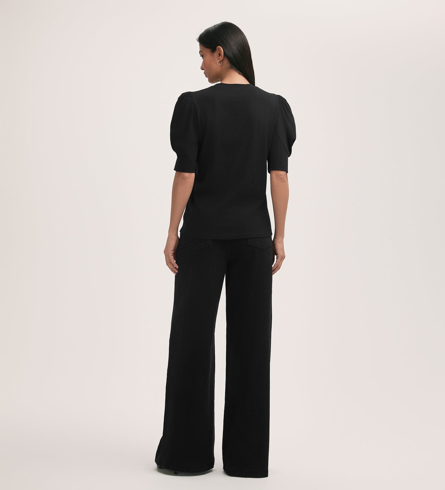 A woman faces away from the camera in an Oriel Black Jersey T-Shirt with a crew neck, short sleeves, and gathered details, paired with wide-leg black pants against a plain light background.