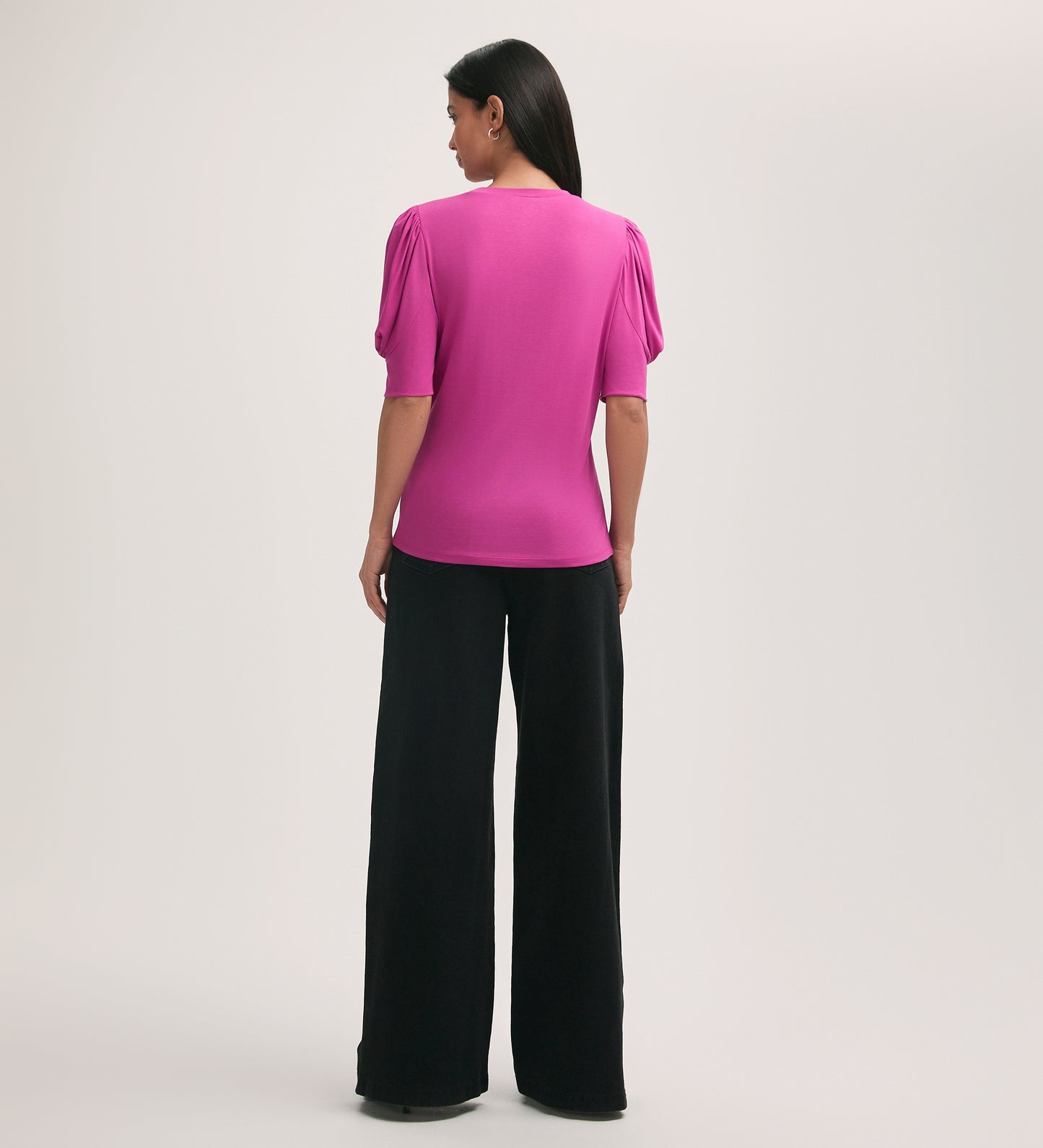 A woman faces away, wearing the Oriel Magenta Jersey T-Shirt with puffed shoulders and a crew neck, made from soft jersey fabric, paired with loose black wide-leg pants against a plain light background.