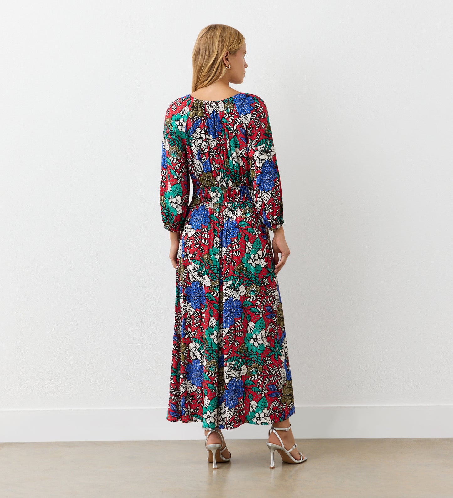 A woman stands indoors facing a white wall, wearing the Piper Red Floral Crepe Midi Tea Dress with a shirred waist and long sleeves. Silver high-heeled sandals complete her look, and her blonde hair falls loosely as the dress reaches her ankles.