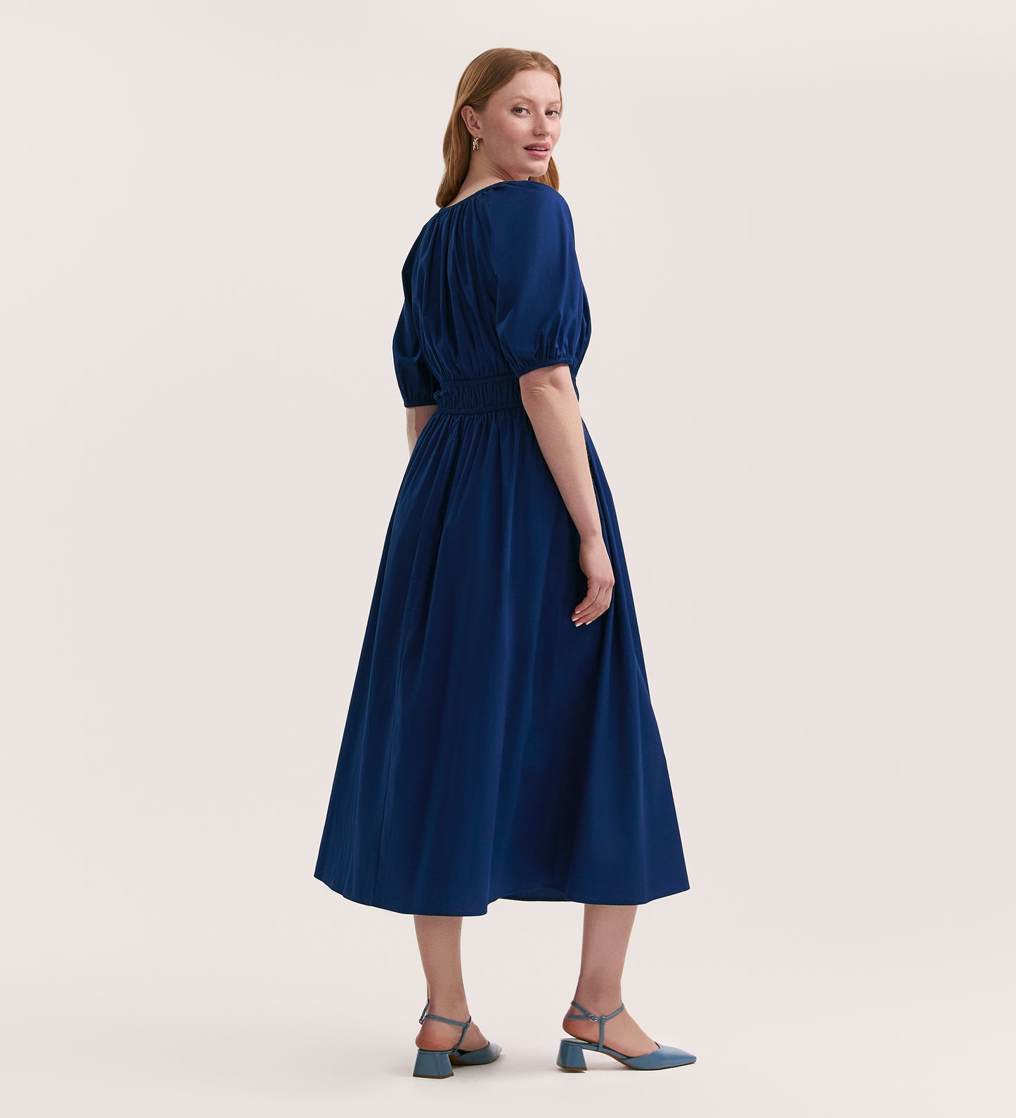 A woman with light skin and long, straight hair stands slightly turned, wearing the Piper Indigo Cotton Midi Smock Dress—a timeless, breathable cotton piece—paired with blue heels against a plain light background.