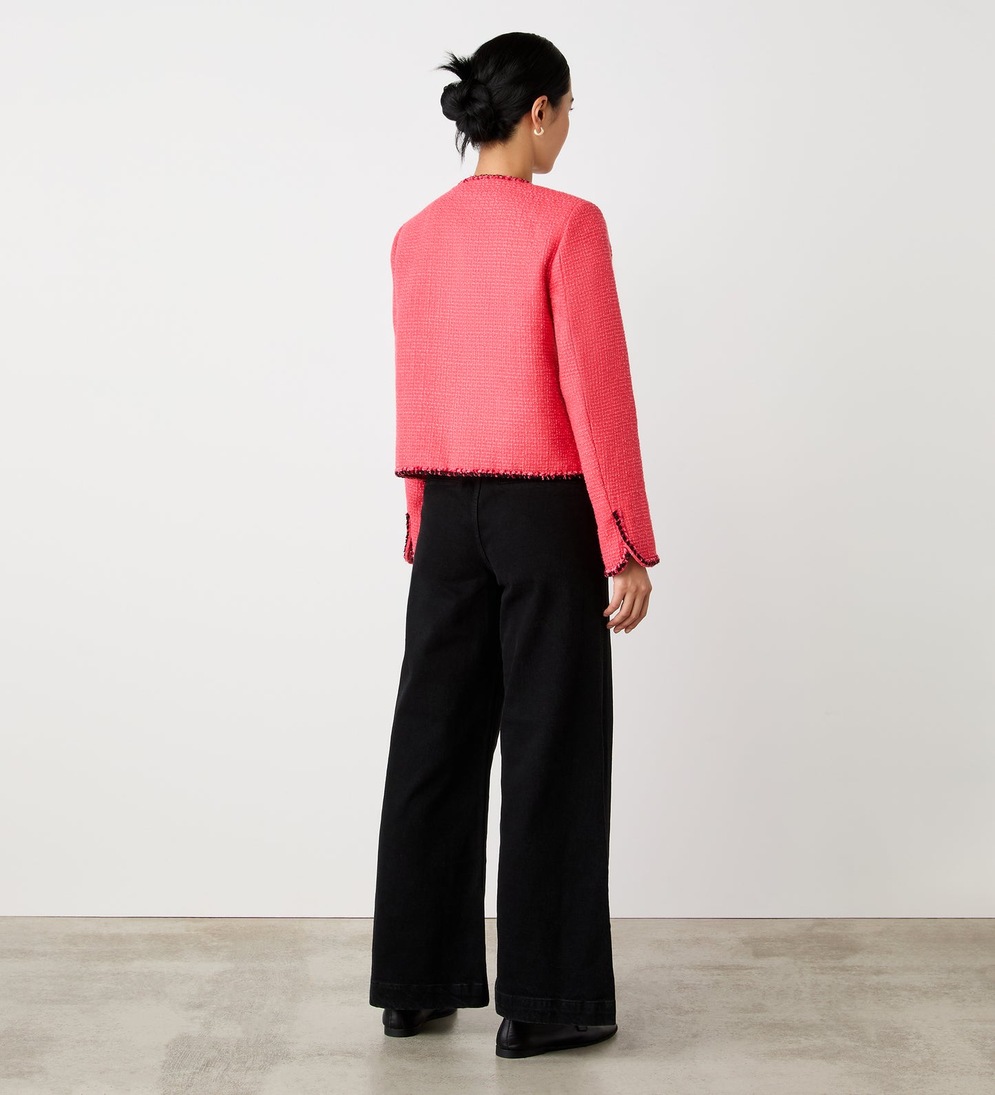 A person with dark hair in a low bun stands facing away, wearing the Polly Coral Jacket—a bright pink decorative tweed cropped style—and wide-leg black pants, on a concrete floor with a plain white wall backdrop.