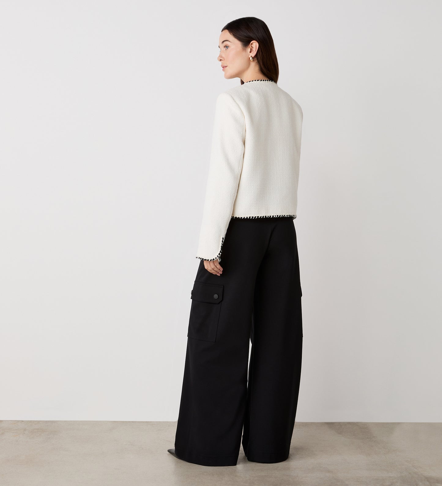 A woman with dark hair stands indoors facing away, wearing the Polly Ivory Jacket—a white tweed piece with contrast trim—and wide-leg black cargo pants, set against a plain light wall and floor.