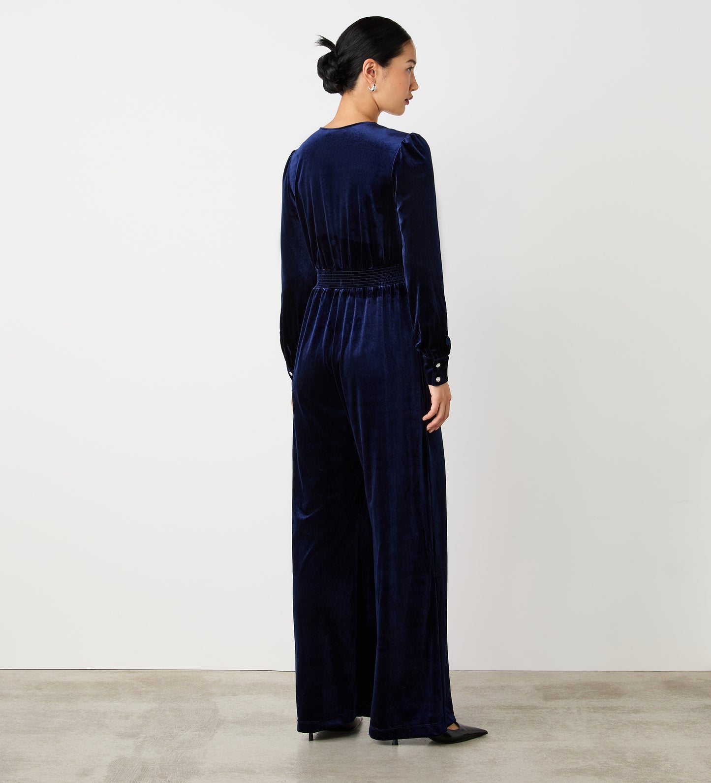 A woman stands indoors, facing away, modeling the Posie Navy Velvet Jumpsuit. The jumpsuit features a flattering fit and wide legs. She pairs it with black pointed-toe heels; her dark hair is styled in a low bun against a white background.