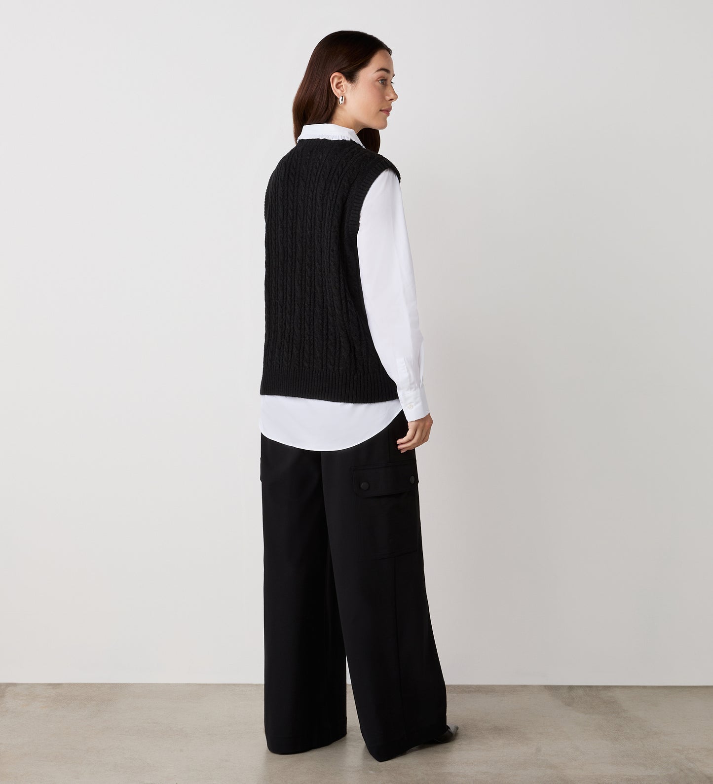 A woman stands facing away, wearing a white button-down shirt under the Primrose Black Crew Neck Knitted Vest and wide-leg black pants with large side pockets, against a plain white wall and concrete floor.