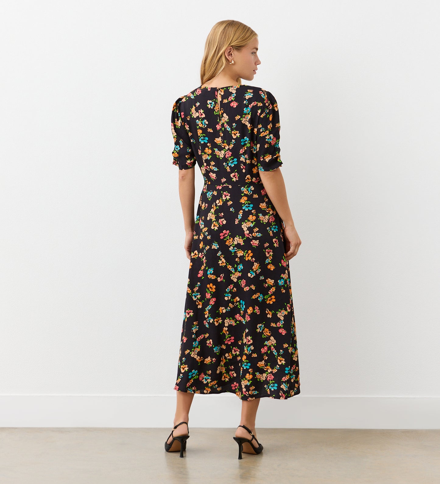 A woman with blonde hair, wearing black heels, stands facing away against a white wall in the Prue Black Floral Crepe Midi Dress featuring colorful floral prints and gathered sleeves.