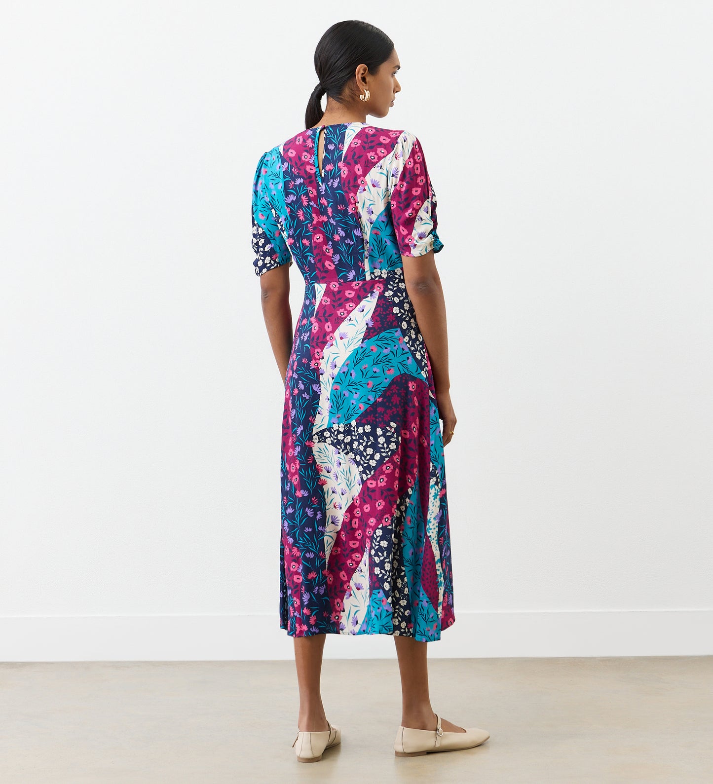 A woman stands indoors, facing away, wearing the Prue Magenta Patchwork Floral Crepe Midi Dress with short gathered sleeves. The dress features blue, pink, white, and purple florals. Her dark hair is tied back and she wears beige flats.