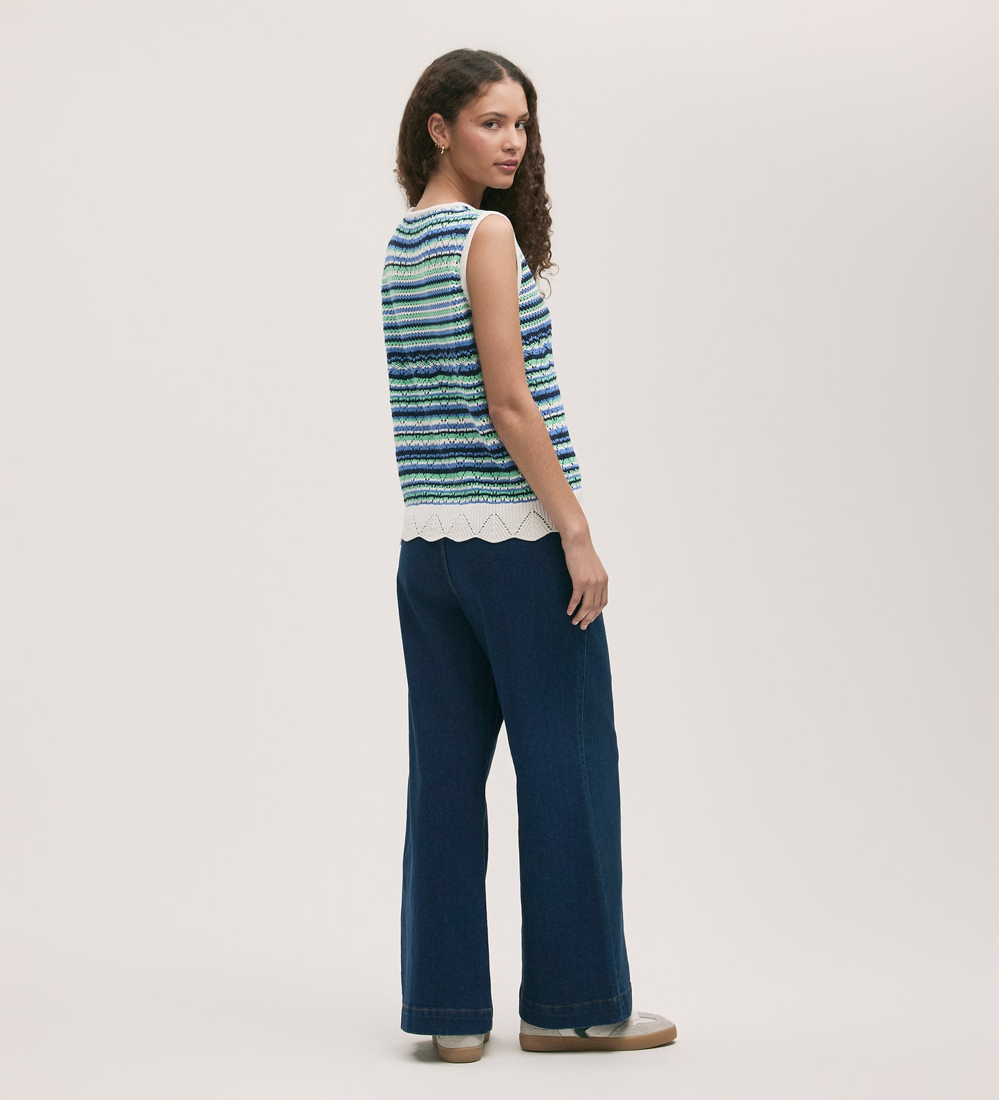 A woman with long curly hair stands sideways, looking over her shoulder. She wears the Porsha Blue Stripe Crochet Vest with a scalloped hem, wide-leg dark blue jeans, and beige shoes against a plain background.