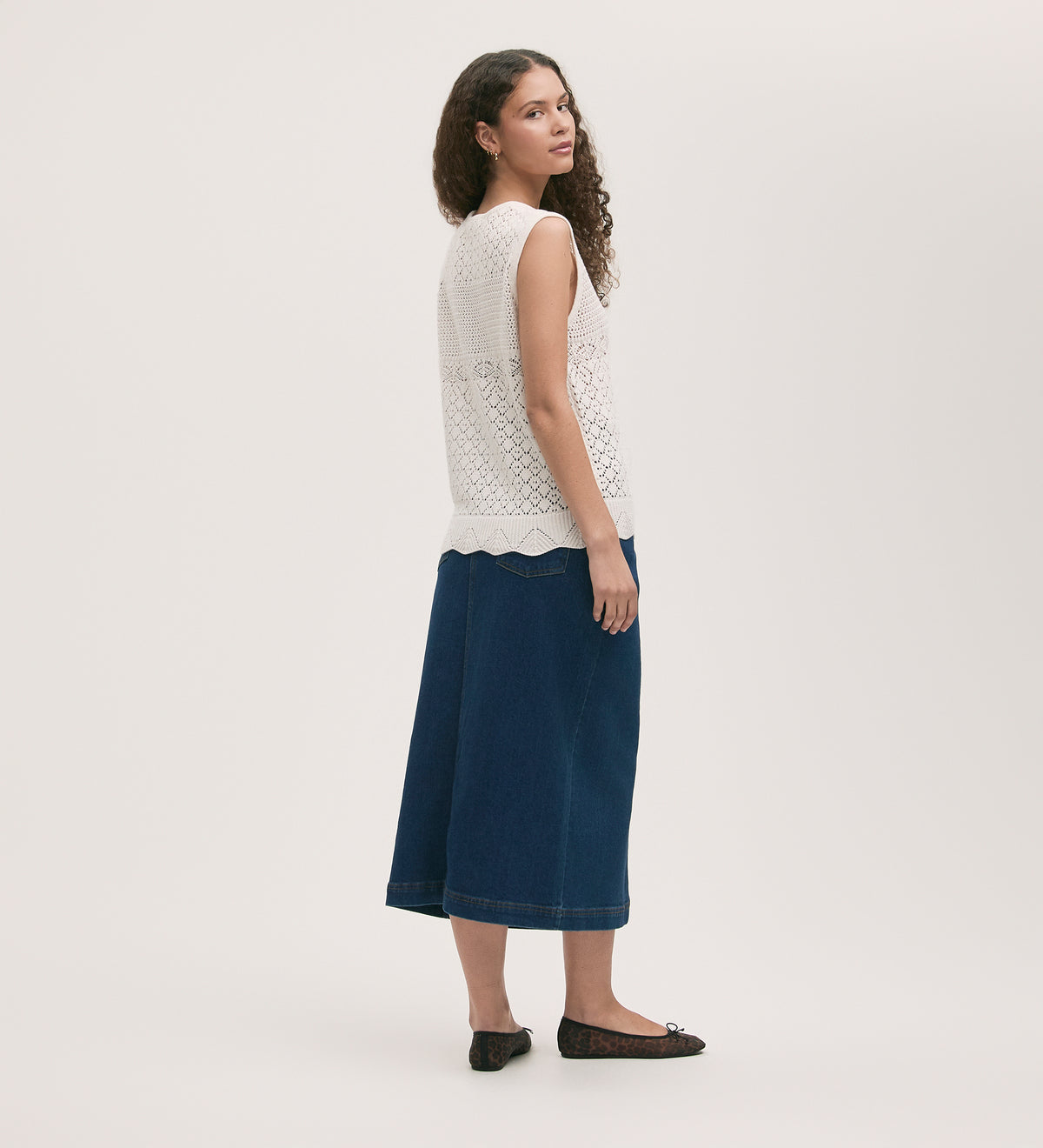 A woman with long curly hair, wearing the Porsha Ivory Crochet Vest, a denim midi skirt, and black flats, stands turned slightly to the side against a plain light background.