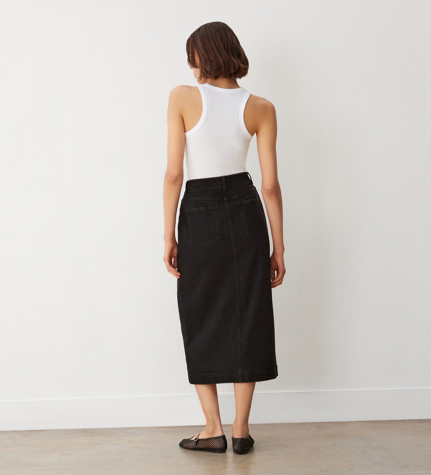 A woman with short brown hair stands facing away against a plain white wall and concrete floor, wearing a white sleeveless tank top, the Rachel Black Cotton Rich Denim Midi Skirt with a straight fit, and black woven flats.
