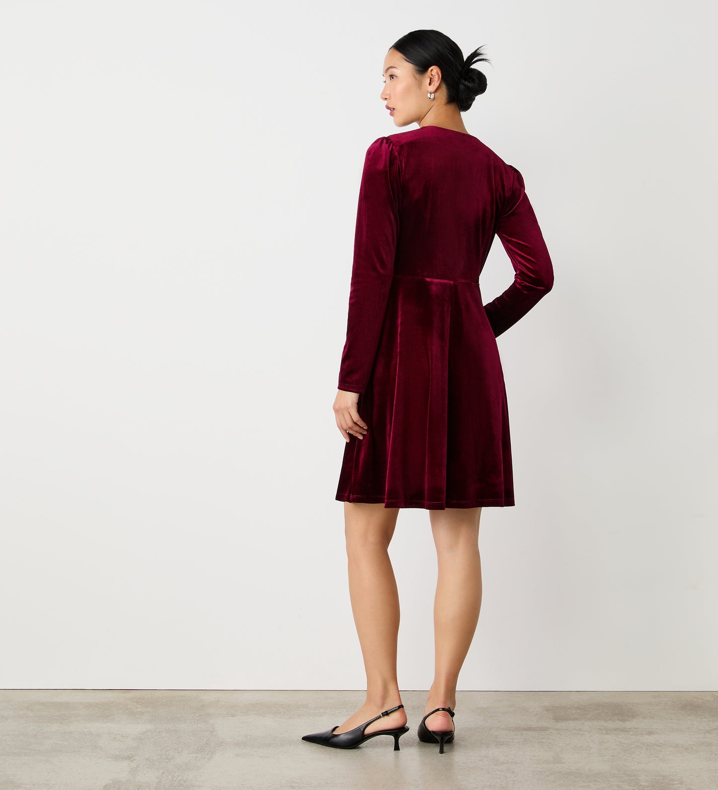 A woman faces away in a knee-length Rae Burgundy Velvet Dress with black slingbacks, her hair in a neat bun. She stands against a white wall on a concrete floor—ideal for any party occasion.