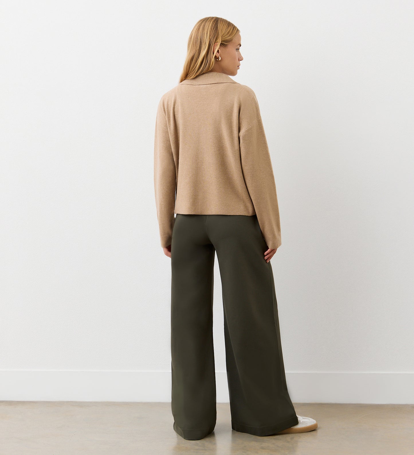A woman faces away from the camera, wearing the Raegan Beige Button Front Cardigan with functional pockets and wide-leg dark olive green pants, set against a plain white wall and light floor.