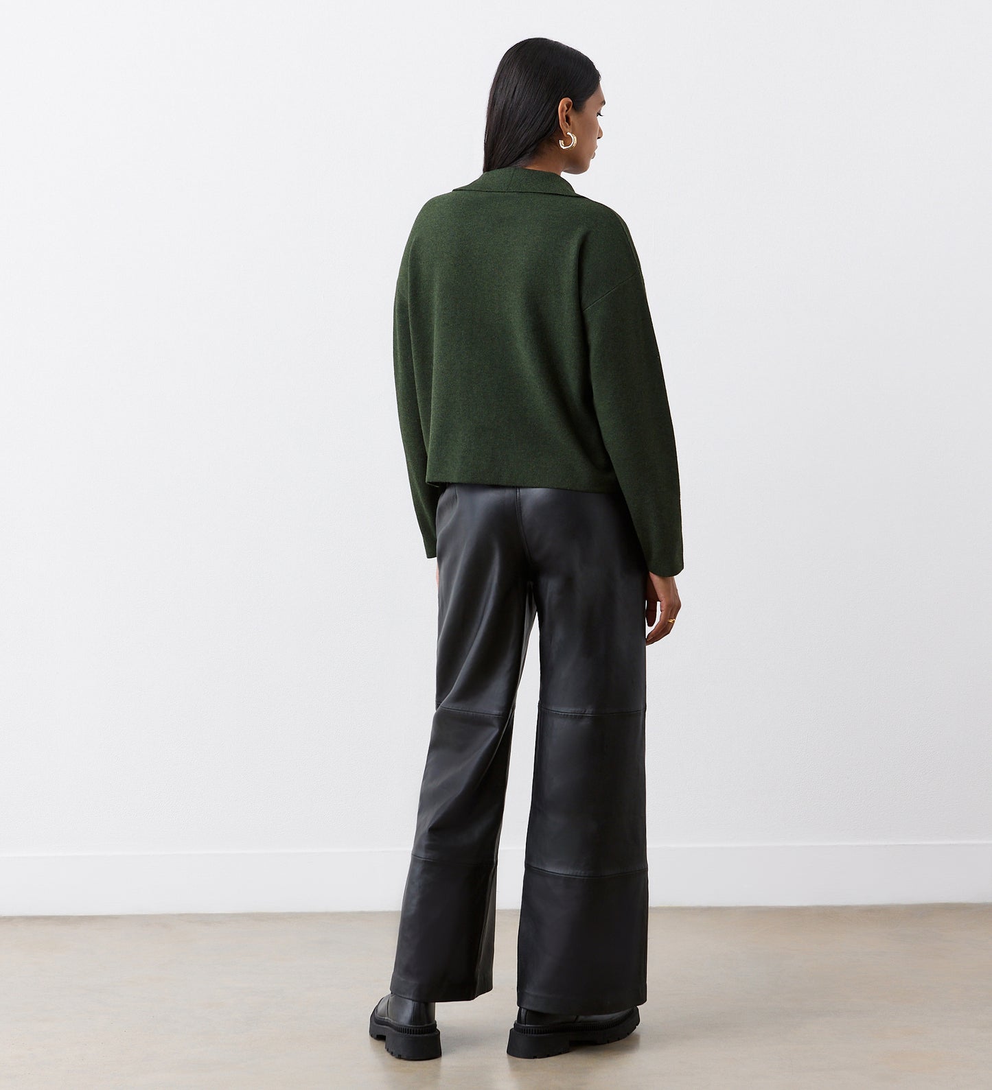 A person stands facing away, wearing a Raegan Green Button Front Cardigan, wide-leg black leather pants, and black shoes, against a plain white wall and light floor.