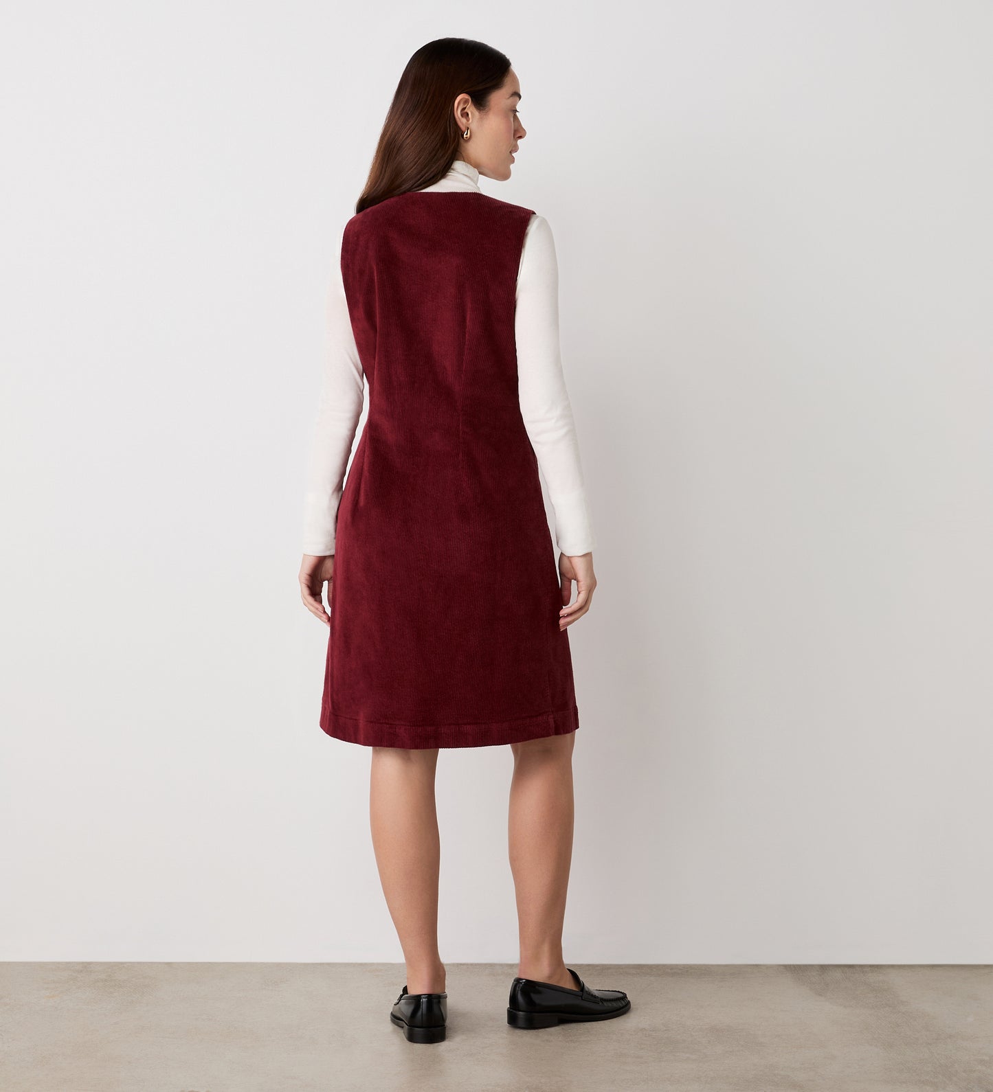 A woman with straight brown hair wears the Reneka Rust Corduroy Knee Length Dress over a long-sleeve white top, paired with black shoes. She stands facing away on a light floor against a plain white background.