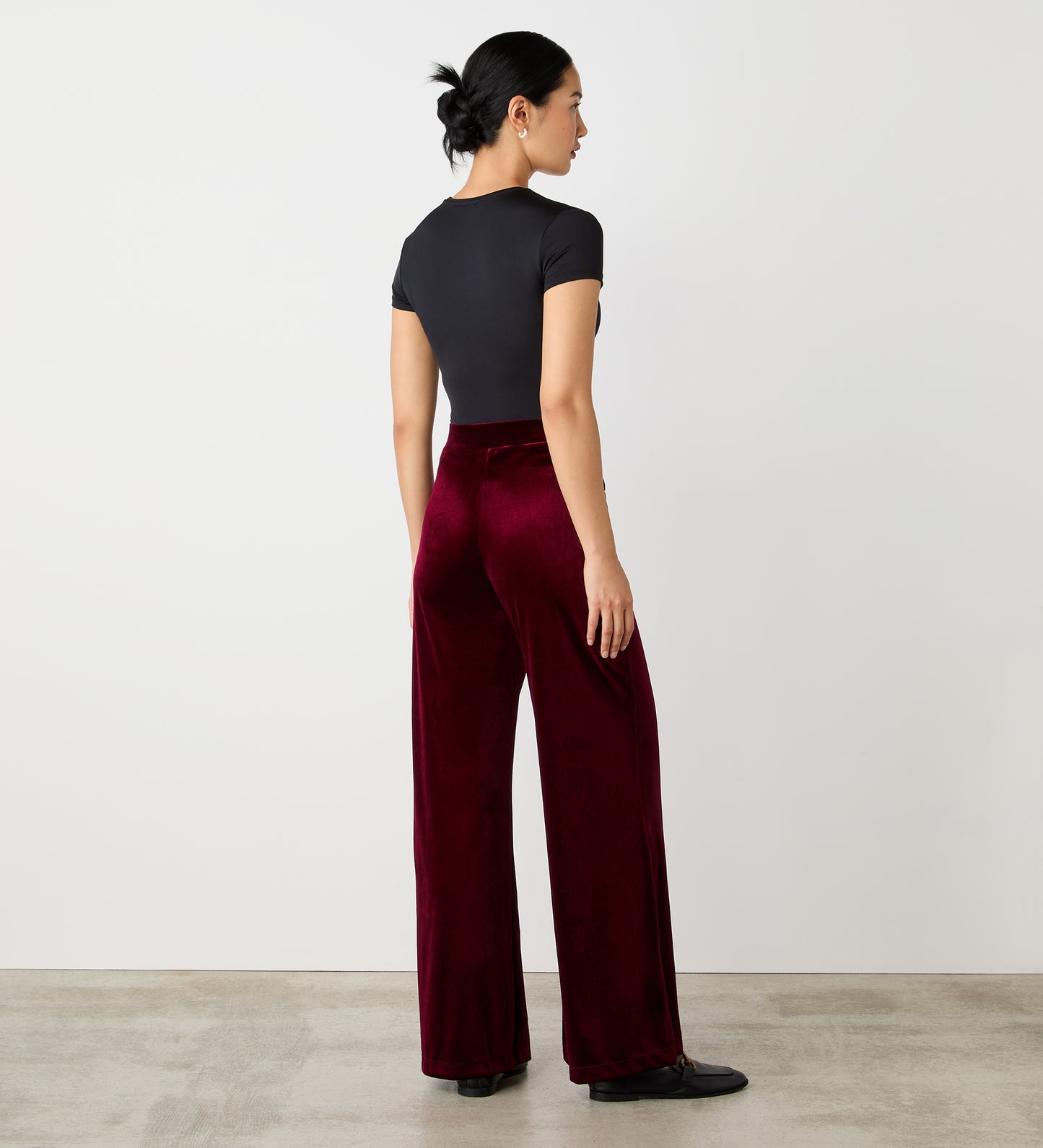 A woman with black hair in a low bun stands on a light floor against a white wall, wearing a black short-sleeve top and high-waisted Reyna Burgundy Velvet Wide Leg Trousers.