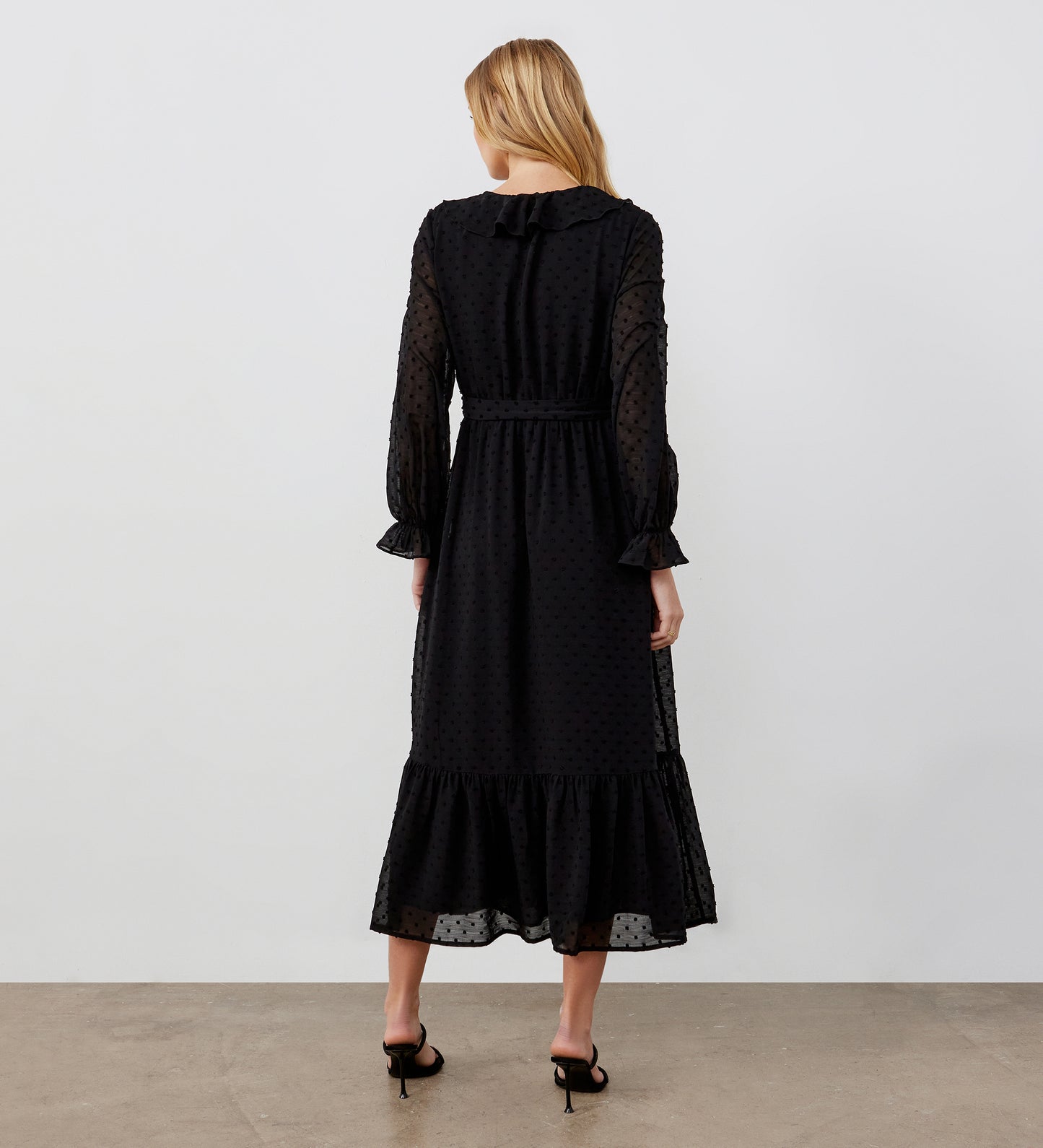 A woman with blonde hair, seen from behind, wears the Rosalie Black Chiffon Midi Wrap Dress with sheer polka-dot fabric and ruffle details, paired with black high-heeled sandals against a plain background.