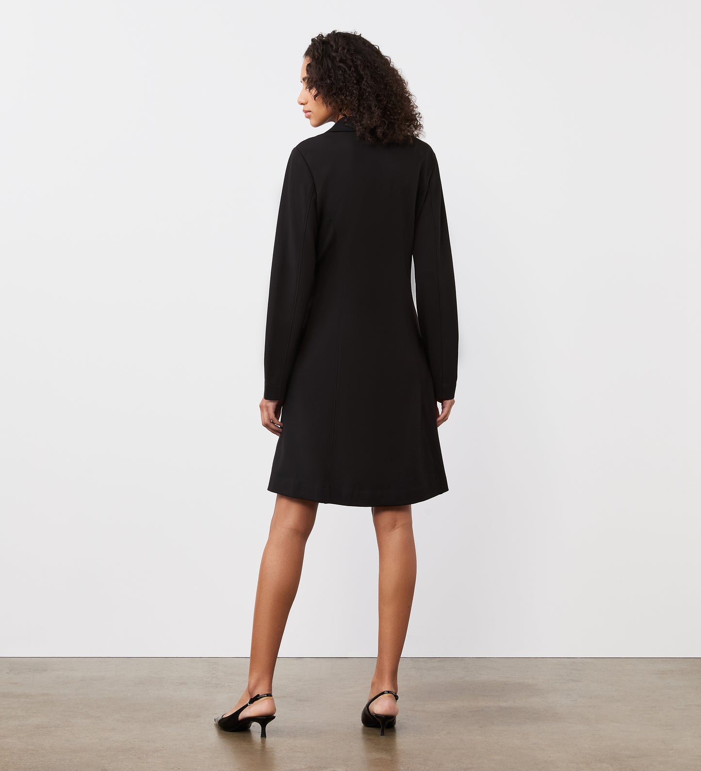 A woman with curly hair stands facing away, wearing the Rosita Black Ponte Jersey Midi Dress and black slingback heels, against a plain white background with a concrete floor.