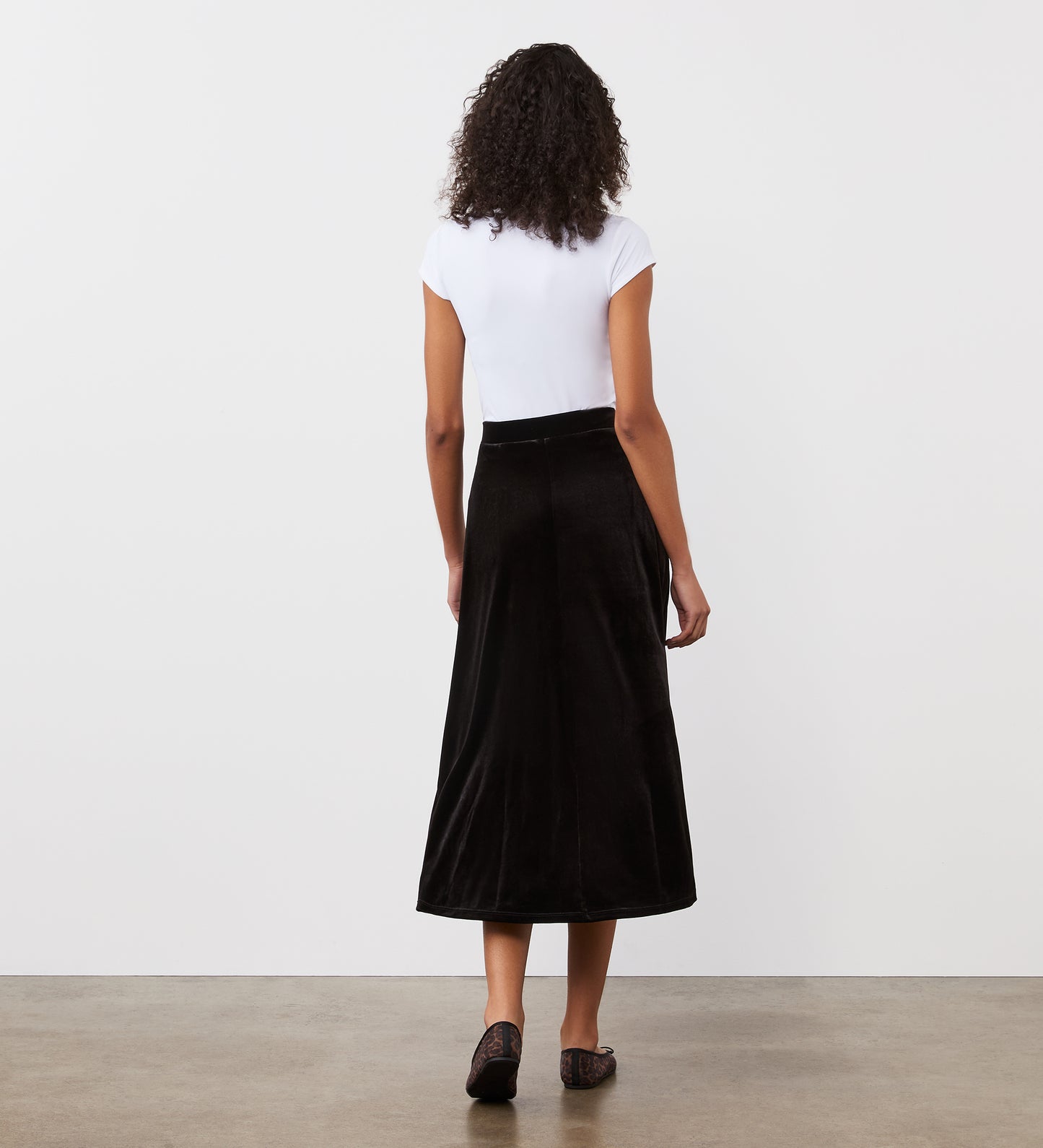 A woman with curly hair, seen from behind, wears a white short-sleeve top and the Ruth Black Velvet Midi Skirt with a thick waistband. She stands on a polished concrete floor against a plain white wall—a versatile wardrobe staple.