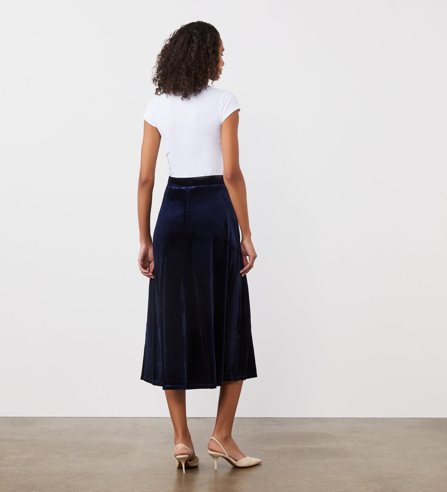 A woman stands indoors facing away, wearing a white short-sleeve top, beige heels, and the Ruth Navy Velvet Midi Skirt against a plain white wall and concrete floor.