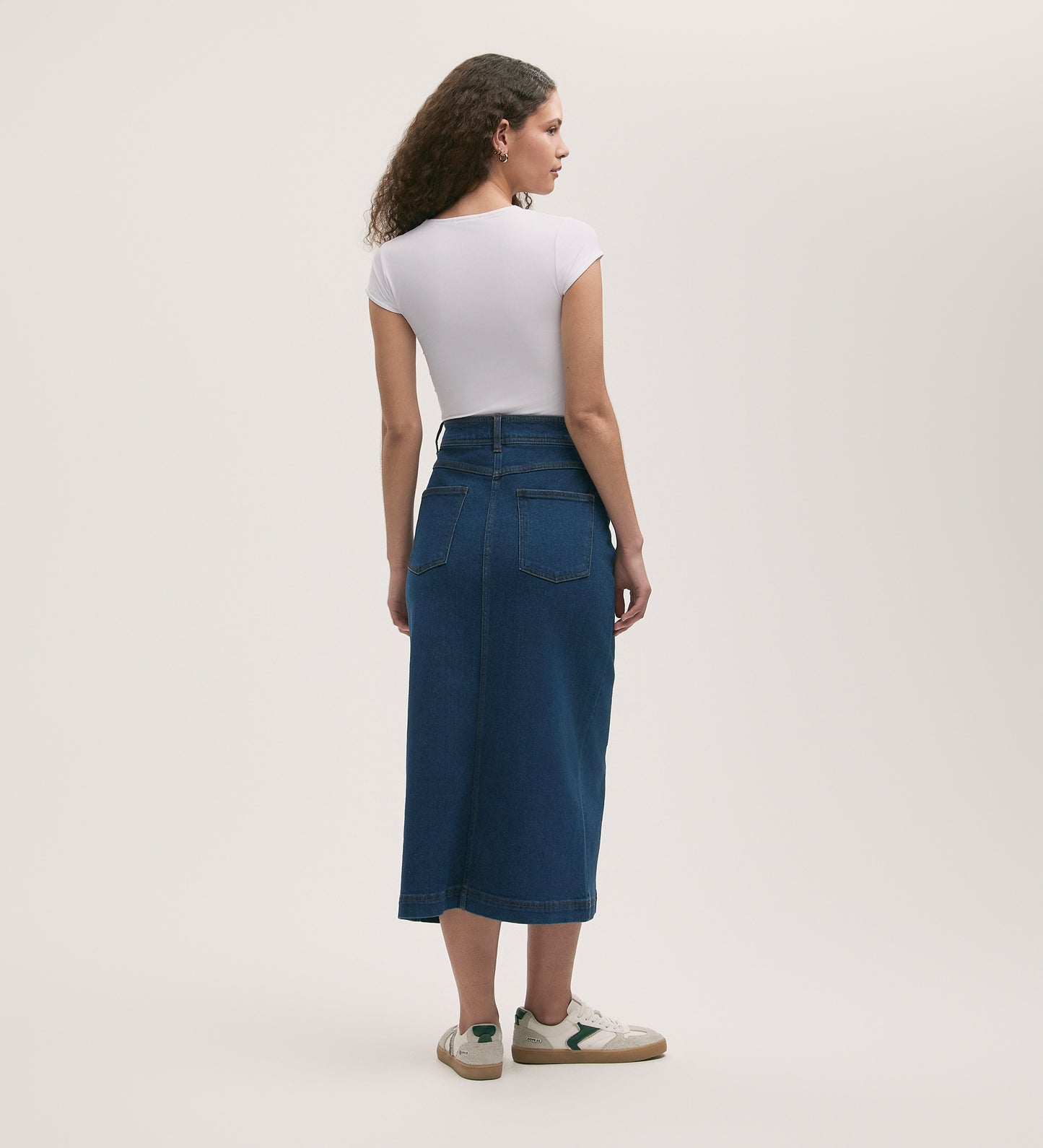 A woman with curly hair, facing away from the camera against a plain background, wears a fitted white short-sleeve top, the Rachel Mid Wash Denim Midi Skirt, and white sneakers.