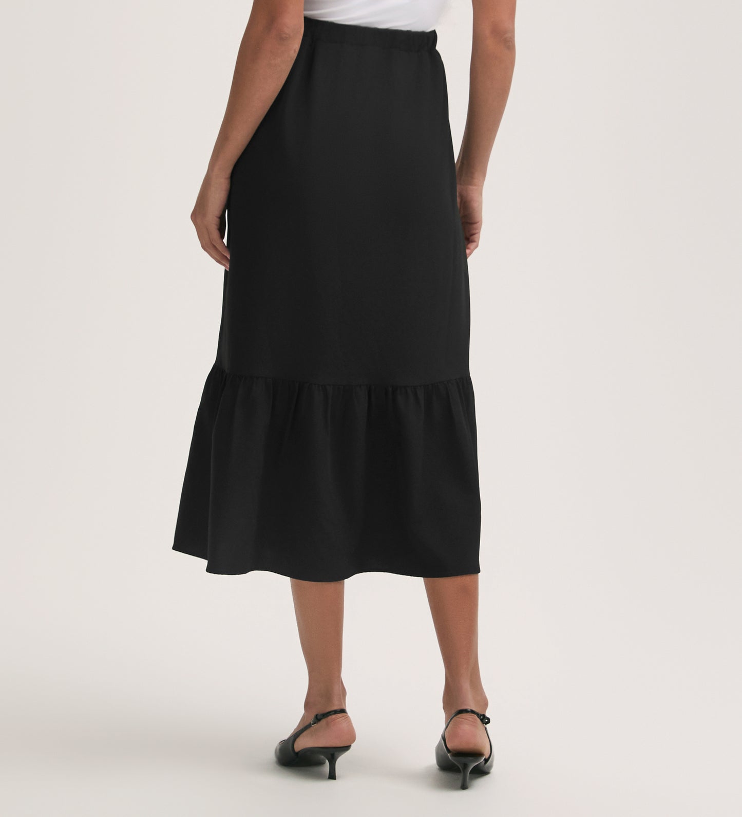 A person is shown from the back wearing the Riley Black Crepe Tiered Midi Skirt, featuring a ruffled hem and elasticated waistband, paired with black heeled sandals against a plain light background.