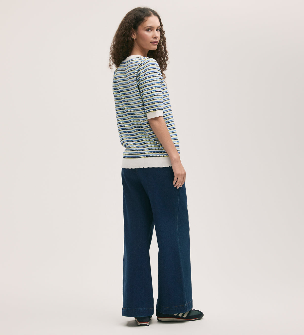 A woman with curly hair stands slightly turned, wearing the Rita Blue Stripe Short Sleeve Top and wide-leg blue jeans. She glances over her shoulder against a plain, light background.