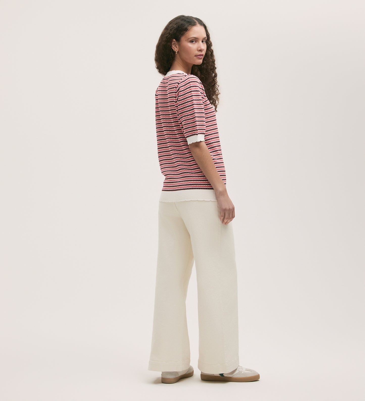 A woman with curly hair stands sideways, looking back at the camera. She wears the Rita Pink Stripe Short Sleeve Top with loose white pants and beige shoes, set against a plain light background.