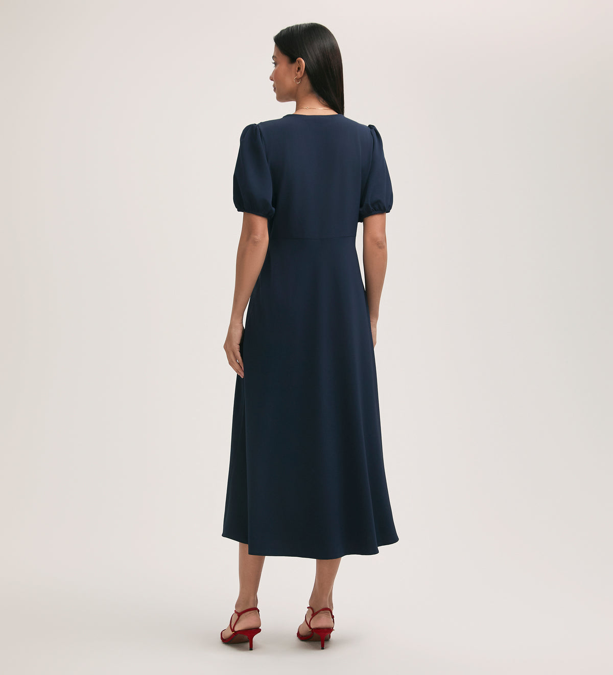 A woman with straight dark hair and red heeled sandals faces away, wearing the Robyn Navy Crepe Midi Tea Dress—crease-free fabric, fitted waist, gently puffed sleeves—against a plain light background.