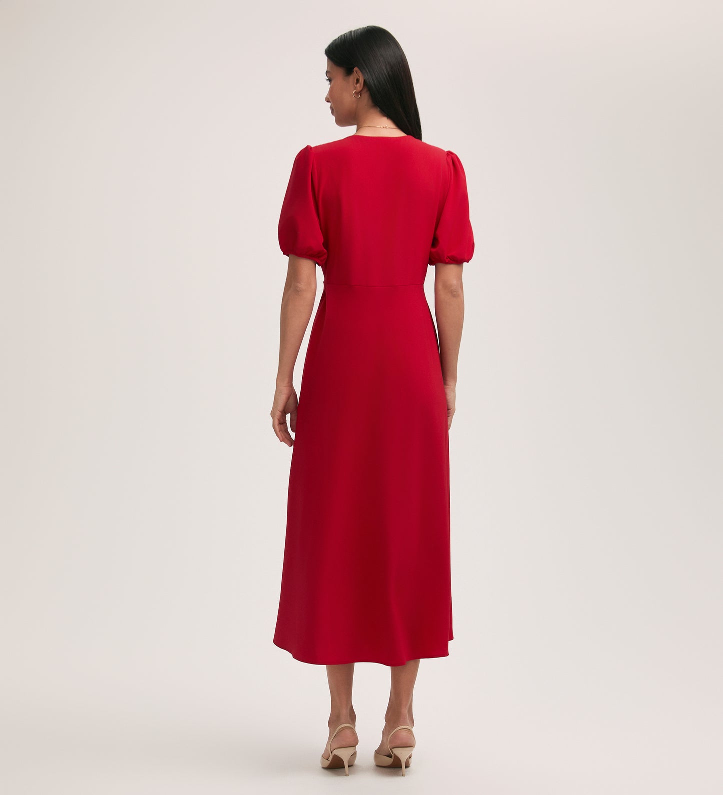 A woman with long dark hair stands facing away in the Robyn Red Crepe Midi Tea Dress, featuring puffed sleeves and a V-neck, paired with beige heeled sandals against a plain light background.