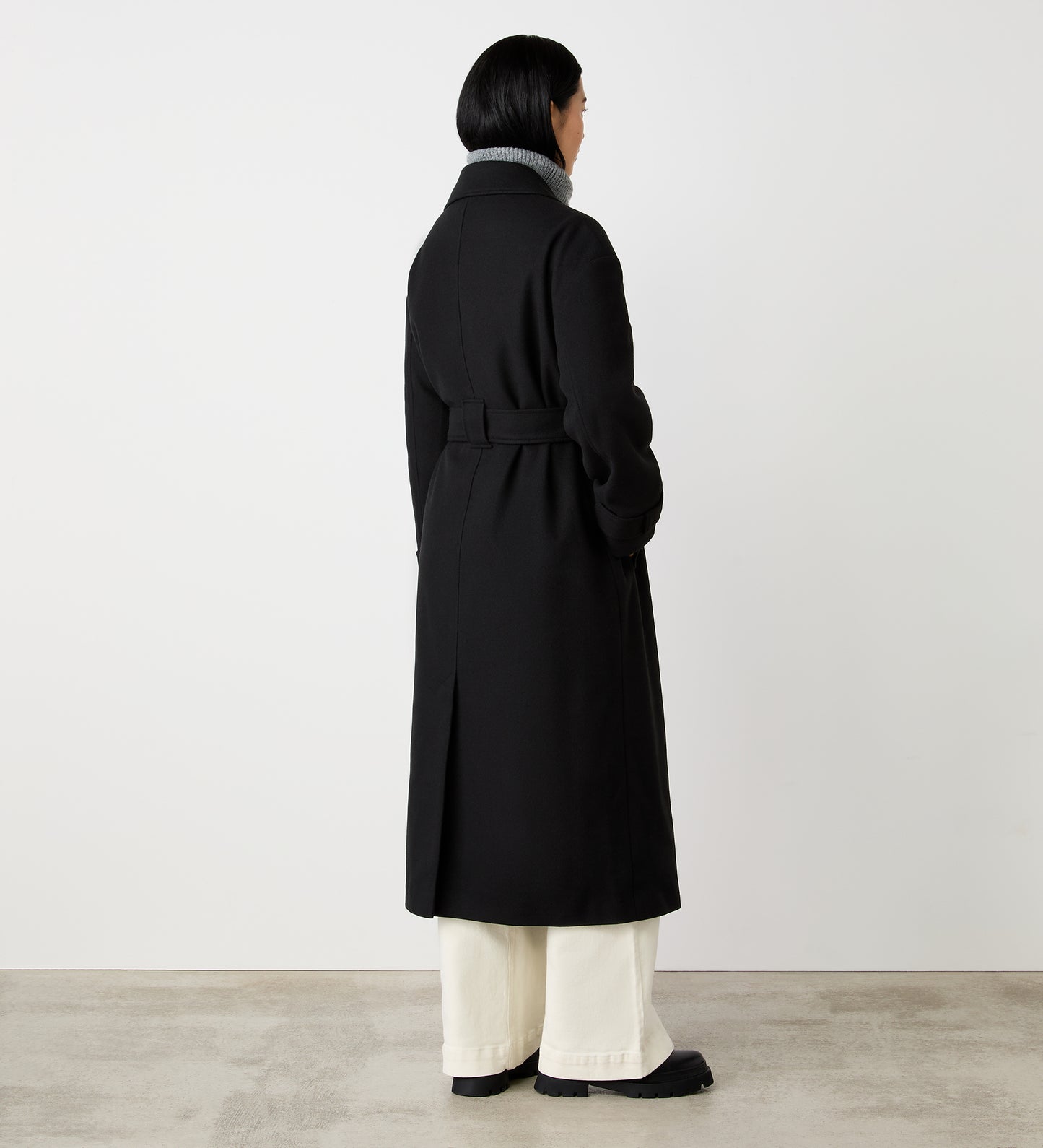 A person with short dark hair faces away, wearing the Sally Black Long Coat with a self-tie belt, paired with light-colored pants and black shoes, standing against a plain white wall and concrete floor.