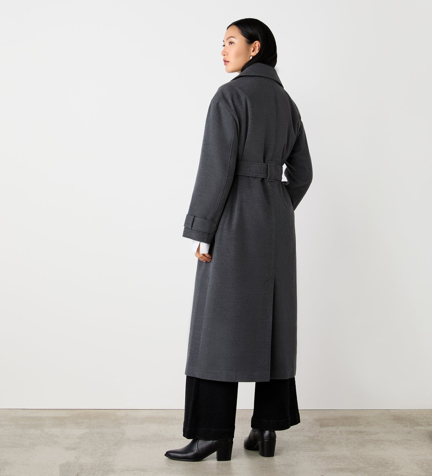 A woman faces away, glancing over her shoulder. She wears the Sally Charcoal Long Coat with a lapel collar and self-tie belt, paired with wide-leg black pants and black heeled boots against a plain white background.