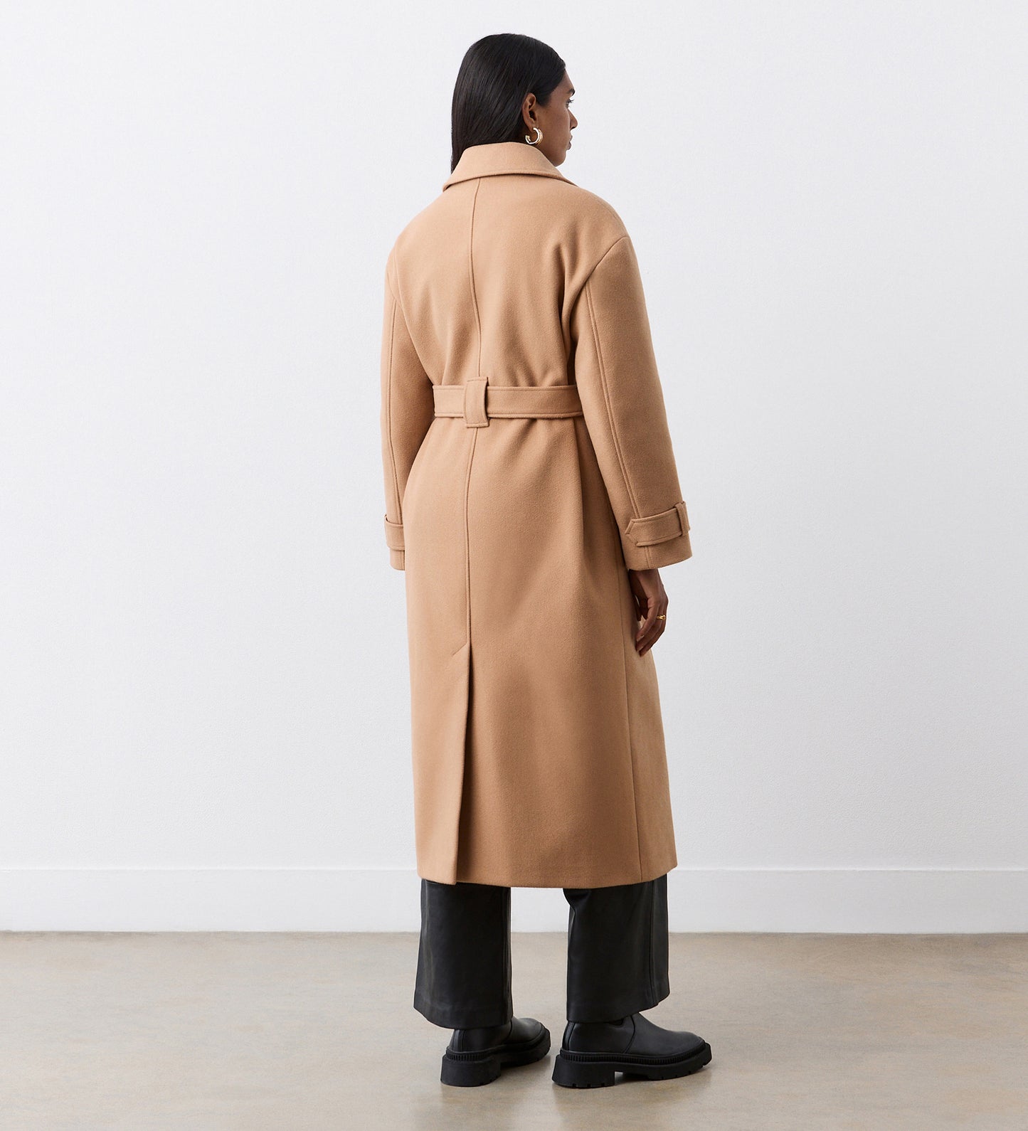 A person stands facing away, wearing the Sally Camel Long Coat with a lapel collar and self-tie belt, paired with black wide-leg pants and black shoes against a plain white wall and concrete floor.