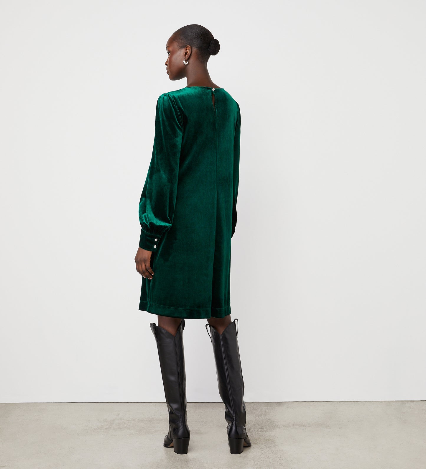 Against a plain white wall and concrete floor, a woman with her hair in a low bun faces away, wearing the Sandrine Bottle Green Velvet Knee Length Dress paired with black knee-high boots.