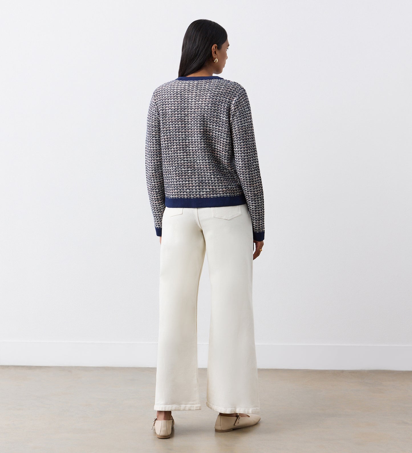 A woman faces away, wearing the Sasha Navy Knitted Cardigan with metallic yarn and gold buttons, paired with ivory wide-leg pants and beige shoes, against a plain white wall and concrete floor.