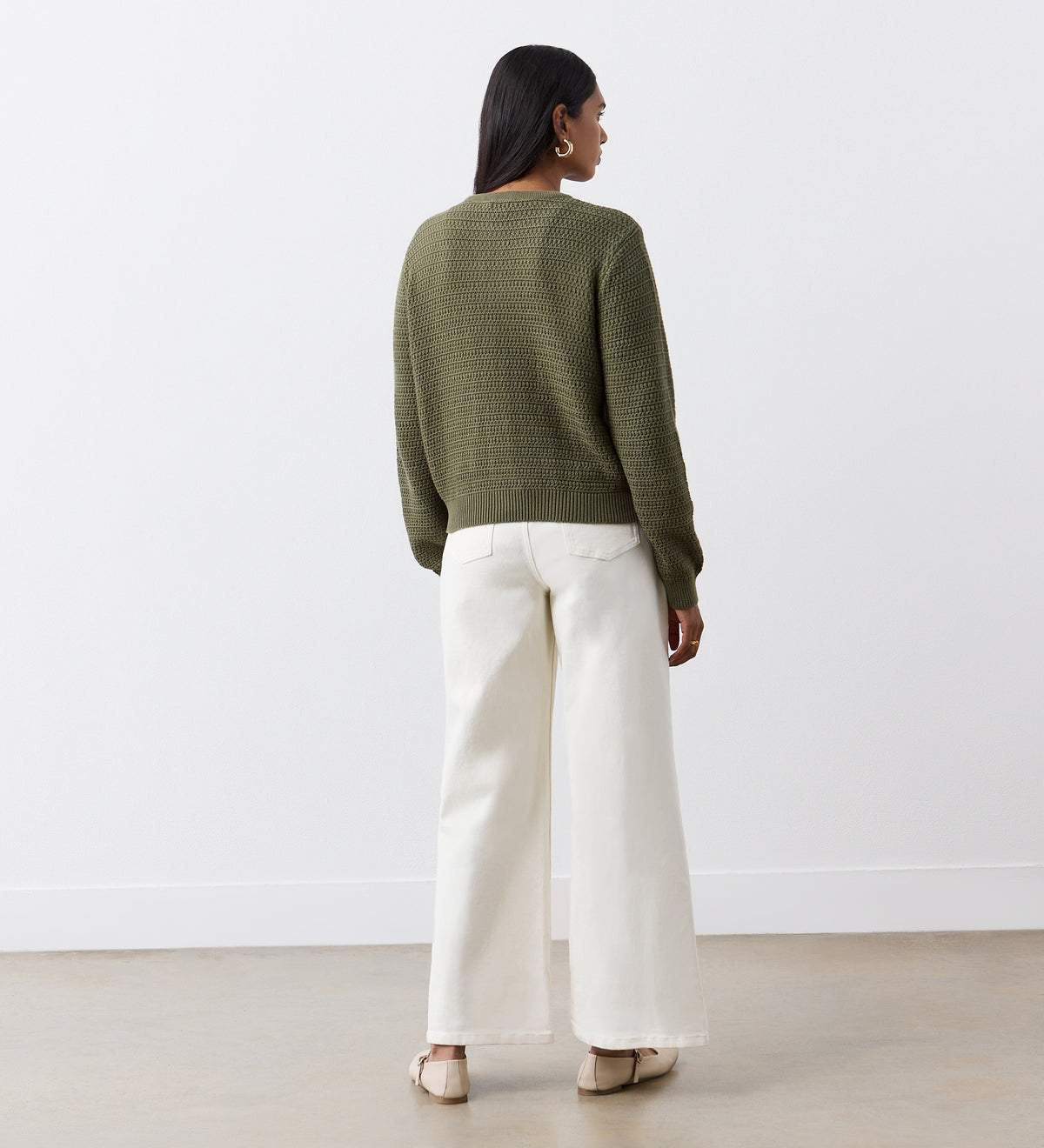 A person with long dark hair faces away, wearing the Saskia Khaki Button Up Cotton Cardigan with gold crest buttons, wide-leg white pants, and beige flats in a minimal indoor space with white walls and a light brown floor.