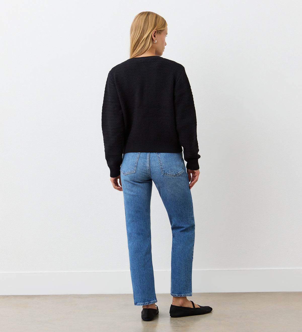 A woman with straight blonde hair stands against a white wall, wearing the Saskia Black Button Up Cotton Cardigan with gold crest buttons, blue jeans, and black flats on a light-colored floor, arms relaxed at her sides.