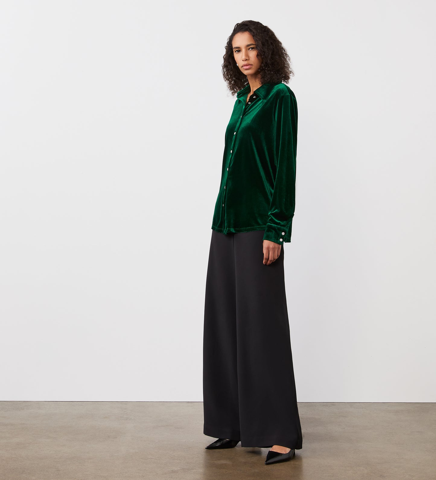 A woman with curly hair wears the Sayge Bottle Green Velvet Shirt, styled with wide-leg black pants, standing against a plain white wall and concrete floor.
