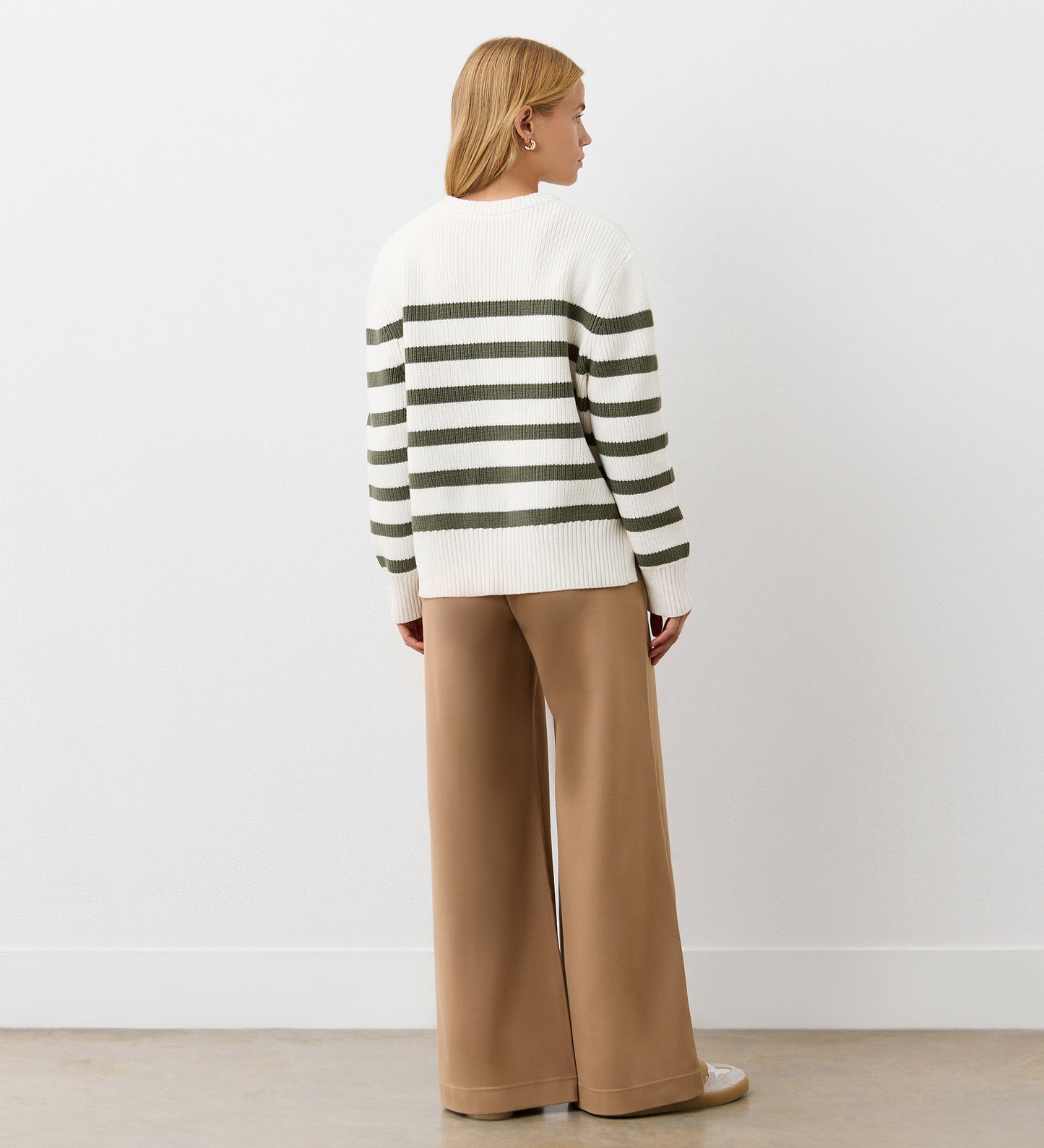 A person with blonde hair faces away, wearing the Serena Khaki Stripe Knitted Sweater—boxy with horizontal olive green stripes and vent sides—paired with wide-leg tan pants, against a plain white wall and light-colored floor.