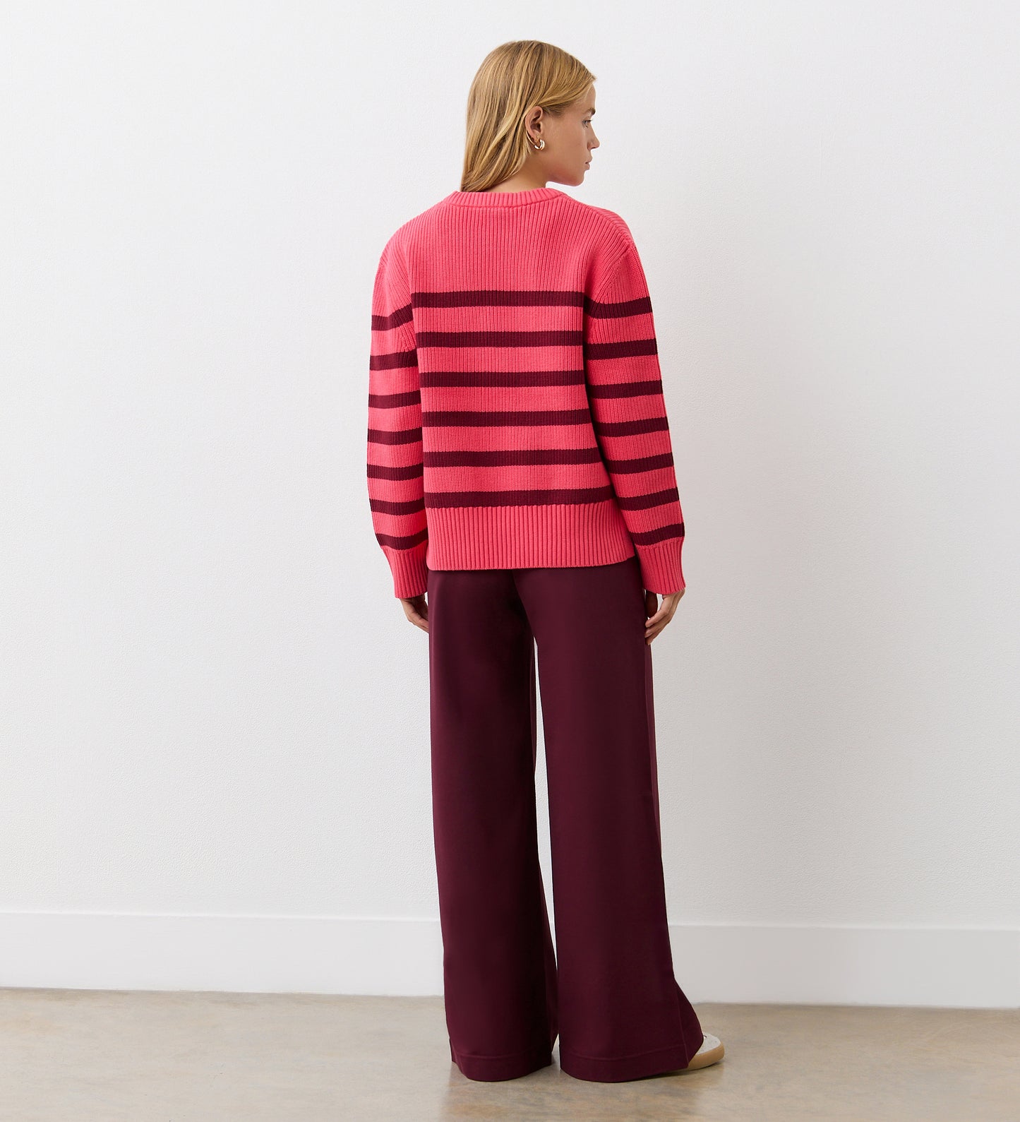A person stands facing away, wearing the Serena Pink Striped Knitted Jumper with dark horizontal stripes and wide-legged maroon pants, set against a plain white wall and light-colored floor.