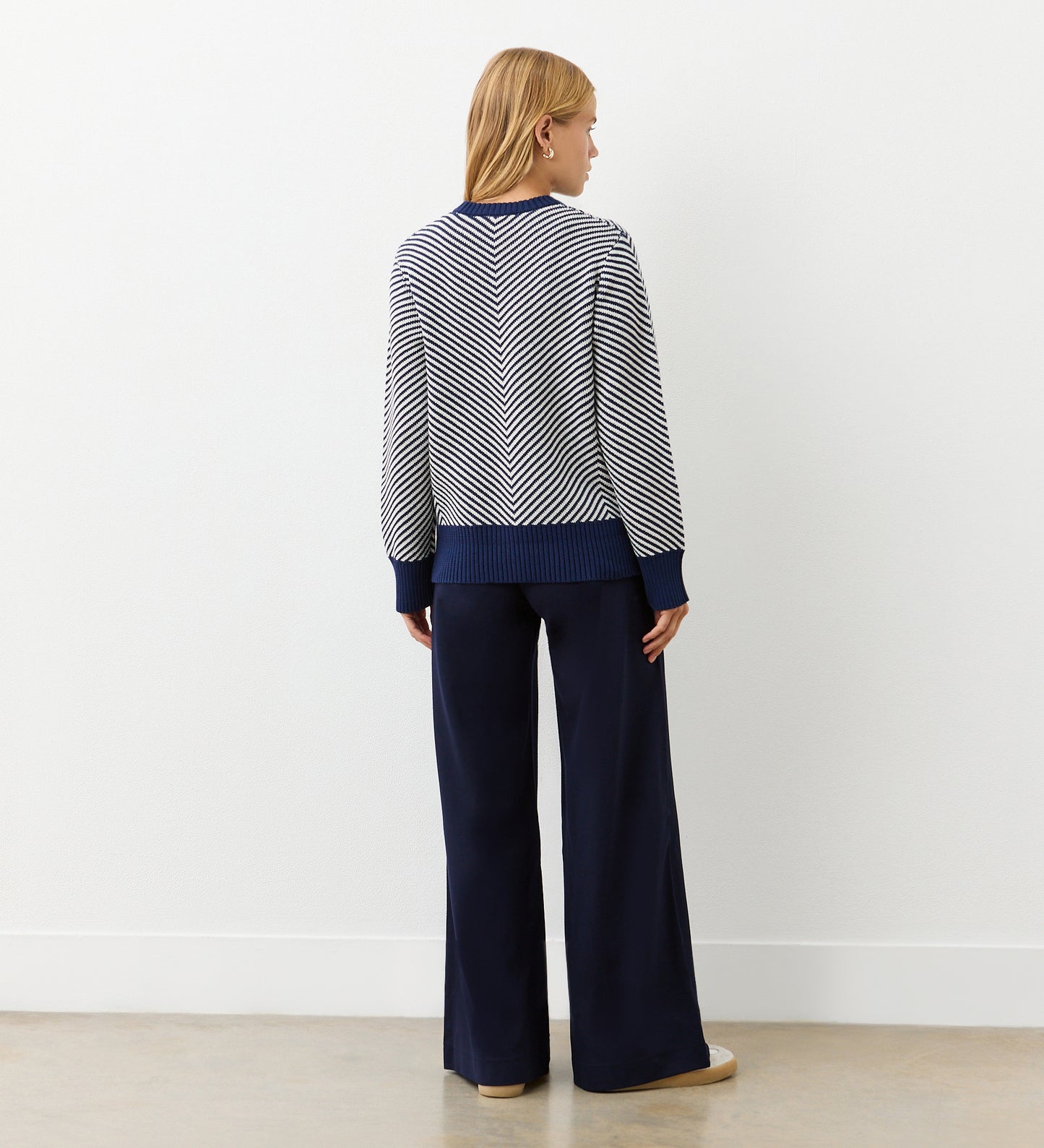 A person with blonde hair faces away, wearing the Stella Navy Chevron Cotton Crew Neck Jumper and loose navy pants, set against a plain light-colored wall and floor background.