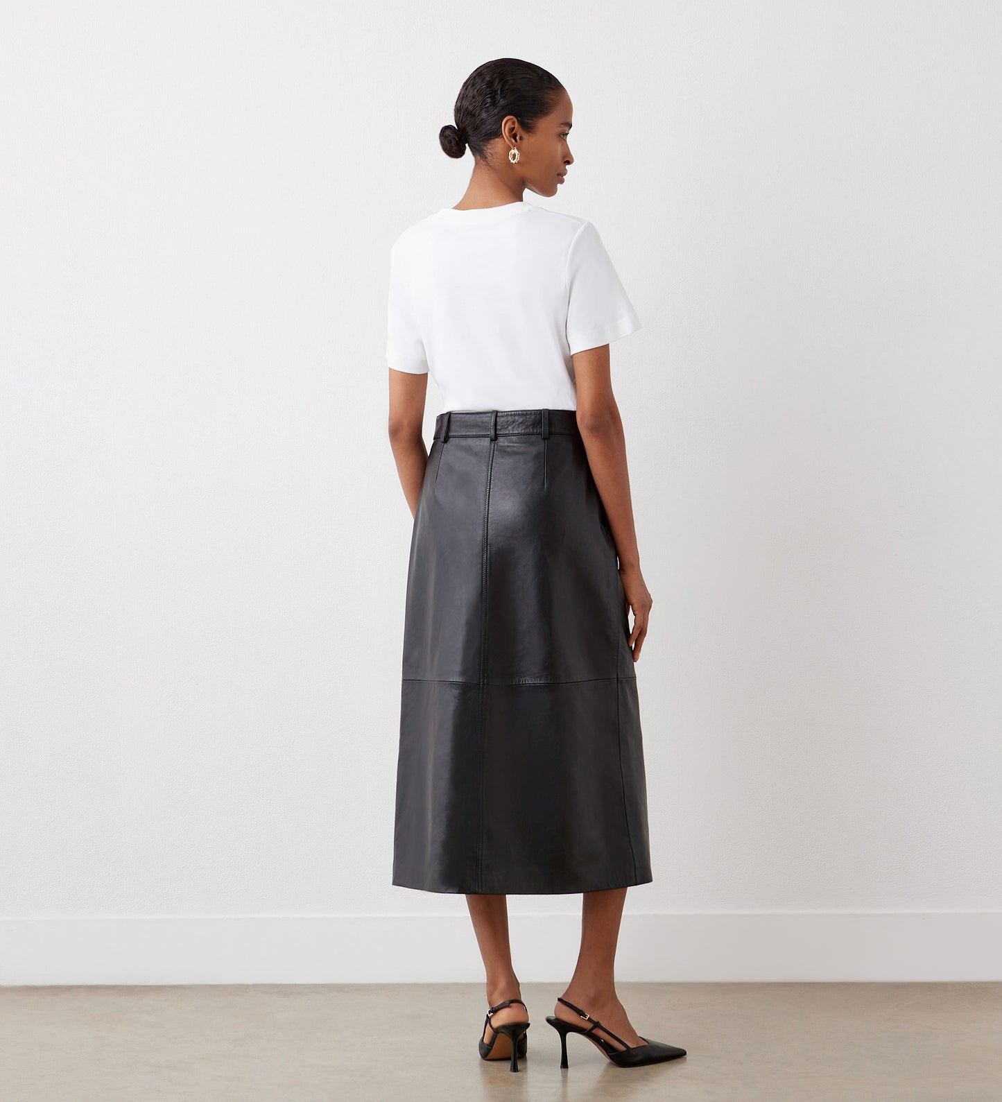 A woman with short hair faces away from the camera, wearing a white short-sleeve top tucked into the Sylvia Black High Waisted Leather Midi Skirt and black heeled sandals, standing against a plain white wall.