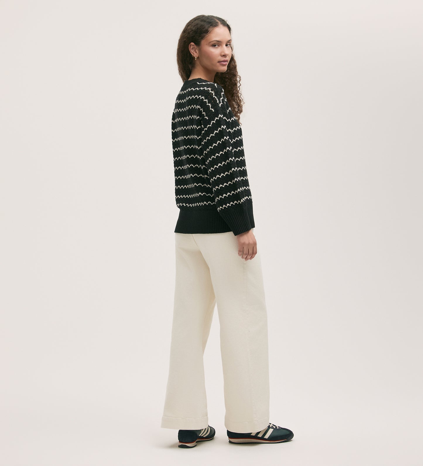 A woman stands sideways, wearing the Sansa Black Crochet Jumper with a black and white zig-zag design, cream wide-leg pants, and black shoes against a plain light background.
