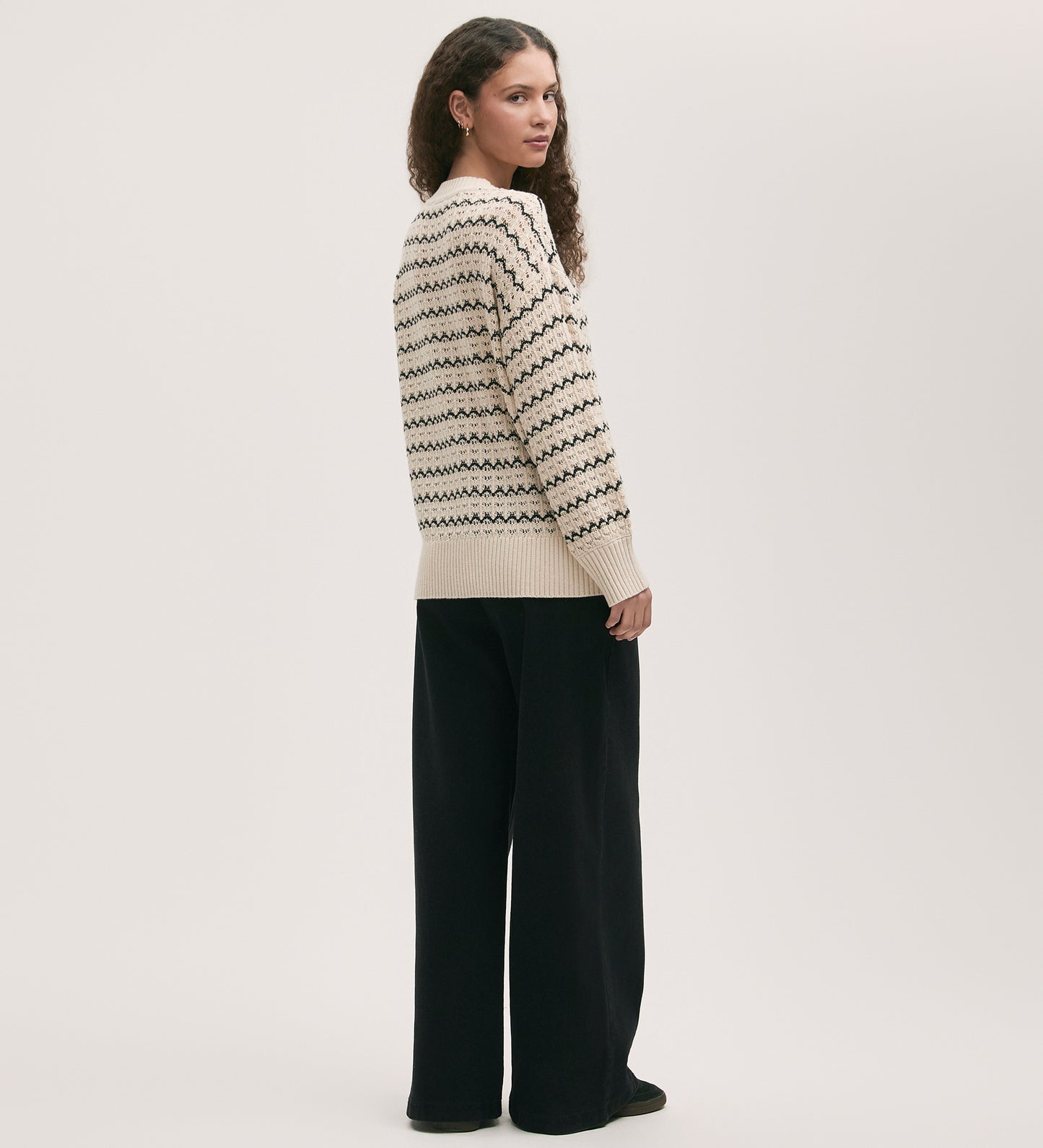 A woman with curly hair stands in profile against a plain light background, looking toward the camera while wearing the Sansa Ivory Crochet Jumper with ribbed detailing and wide-leg black pants.