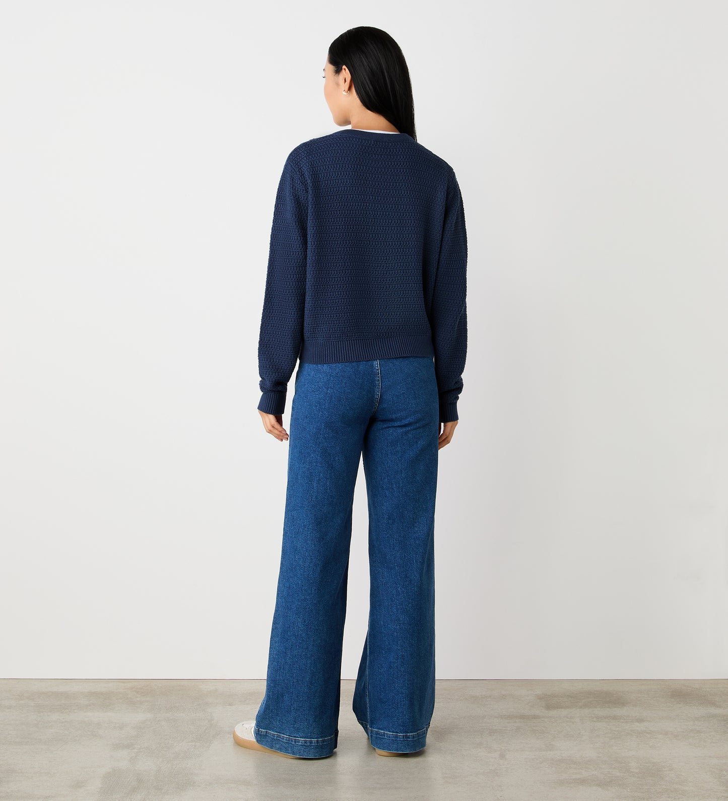 A person with long dark hair stands facing away, wearing the Saskia Dark Navy Button Up Cotton Cardigan and wide-leg blue jeans, against a plain white wall and concrete floor.