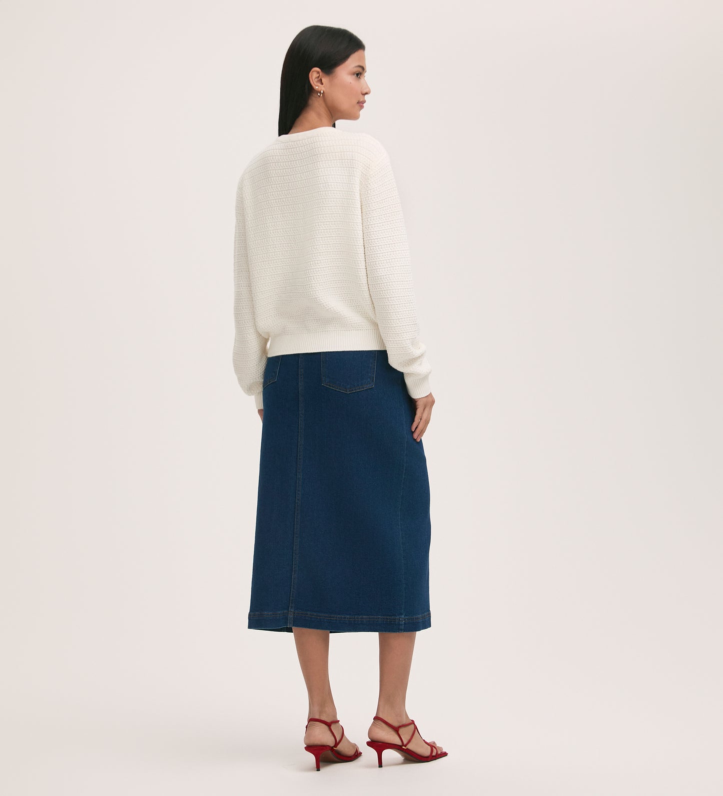 A woman stands facing away against a plain light background, wearing the Saskia Ivory Button Up Cotton Cardigan, a blue denim midi skirt, and red heeled sandals.