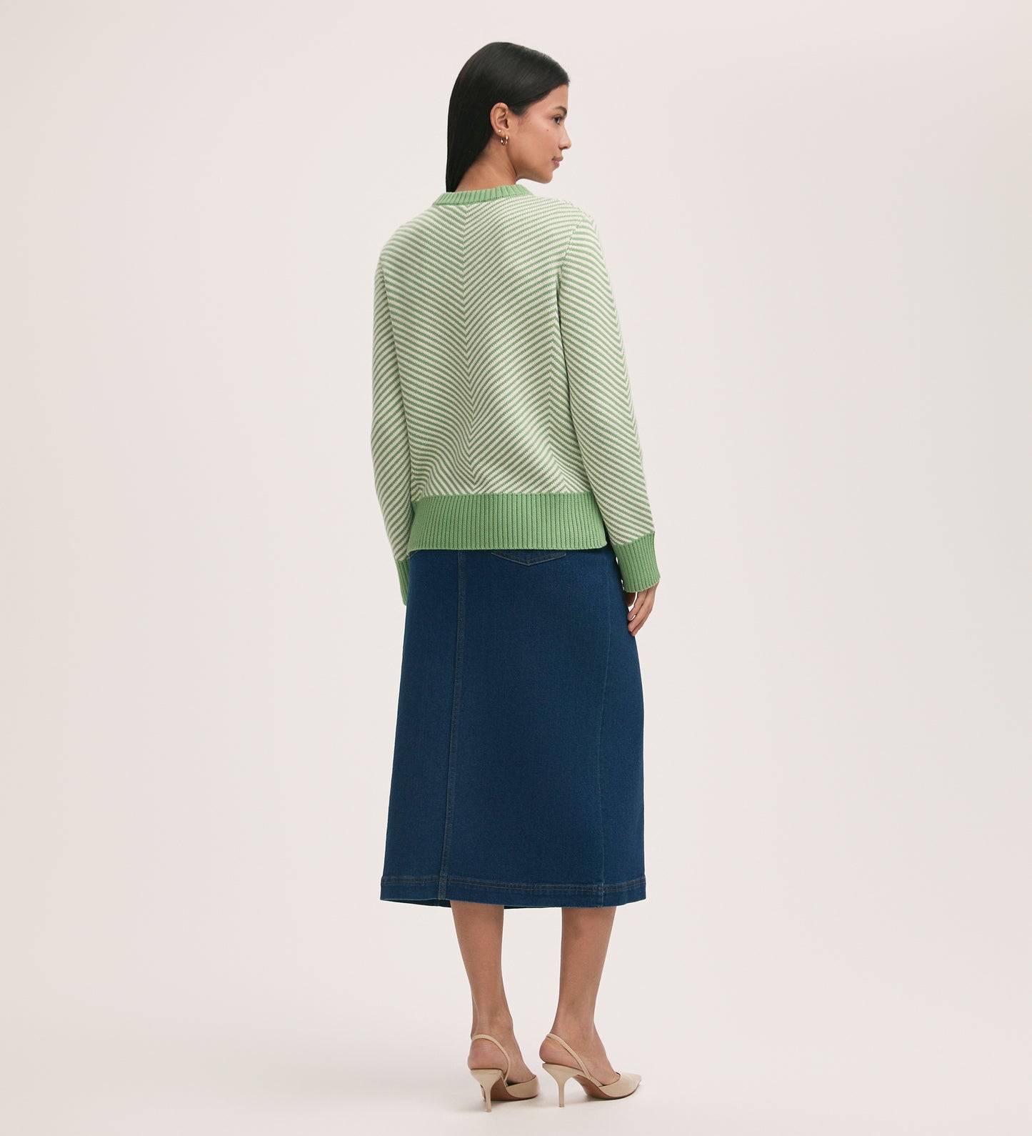 A woman stands facing away, wearing the Stella Green Chevron Cotton Crew Neck Jumper with a dark blue midi denim skirt and beige heeled sandals, set against a plain light background.