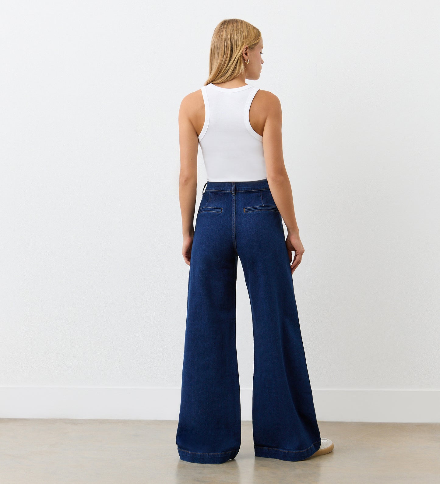 A woman stands facing away in a white sleeveless tank top and Tally Mid Wash Denim Jeans, styled 1970s, in a minimal room with a plain wall and light flooring.