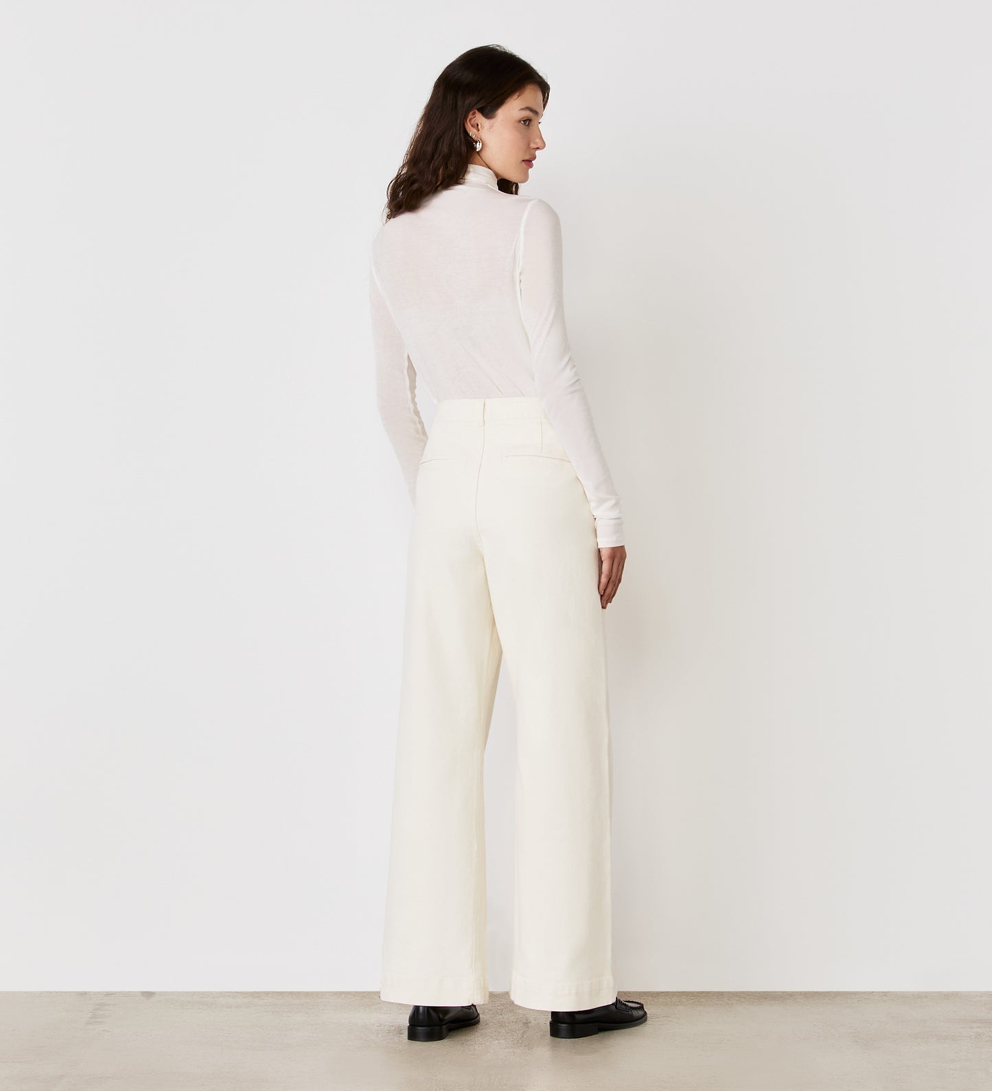 A woman stands indoors facing away, wearing a long-sleeve white turtleneck top, Tally Ecru Denim Jeans with a high waist and wide-leg cut, and black shoes against a plain white background.