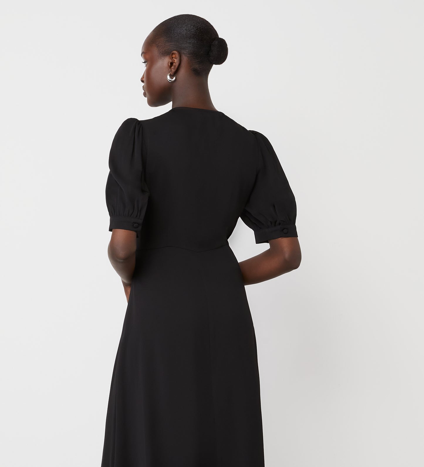 A woman with short hair wears silver hoop earrings and the Tara Black Crepe Midi Dress, featuring puff sleeves and a v-neckline, as she stands facing away from the camera against a plain white background.