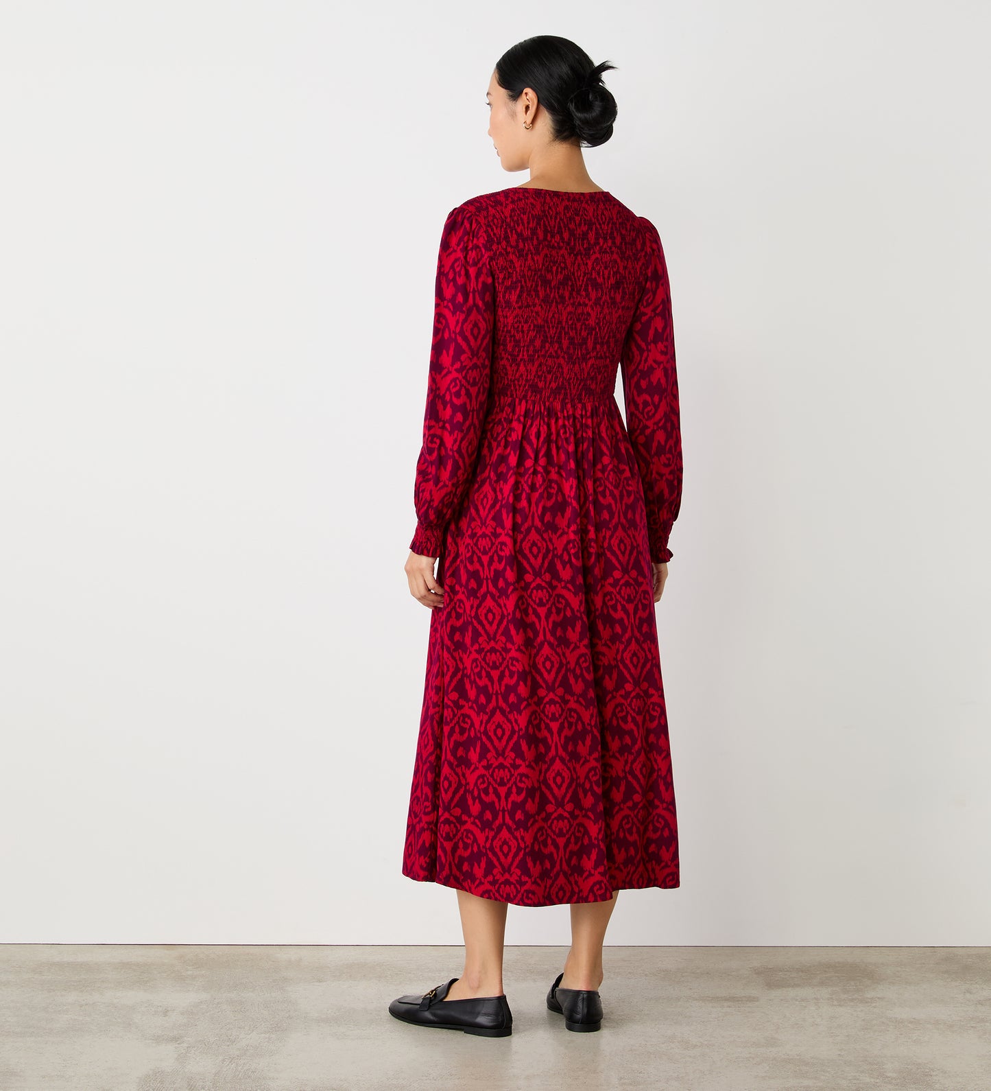 A woman with dark hair tied back wears the Tasmin Pink Ikat Midi Dress, featuring long sleeves and a pink ikat pattern. She pairs it with black flat shoes and stands on a light floor against a plain white wall, facing away.
