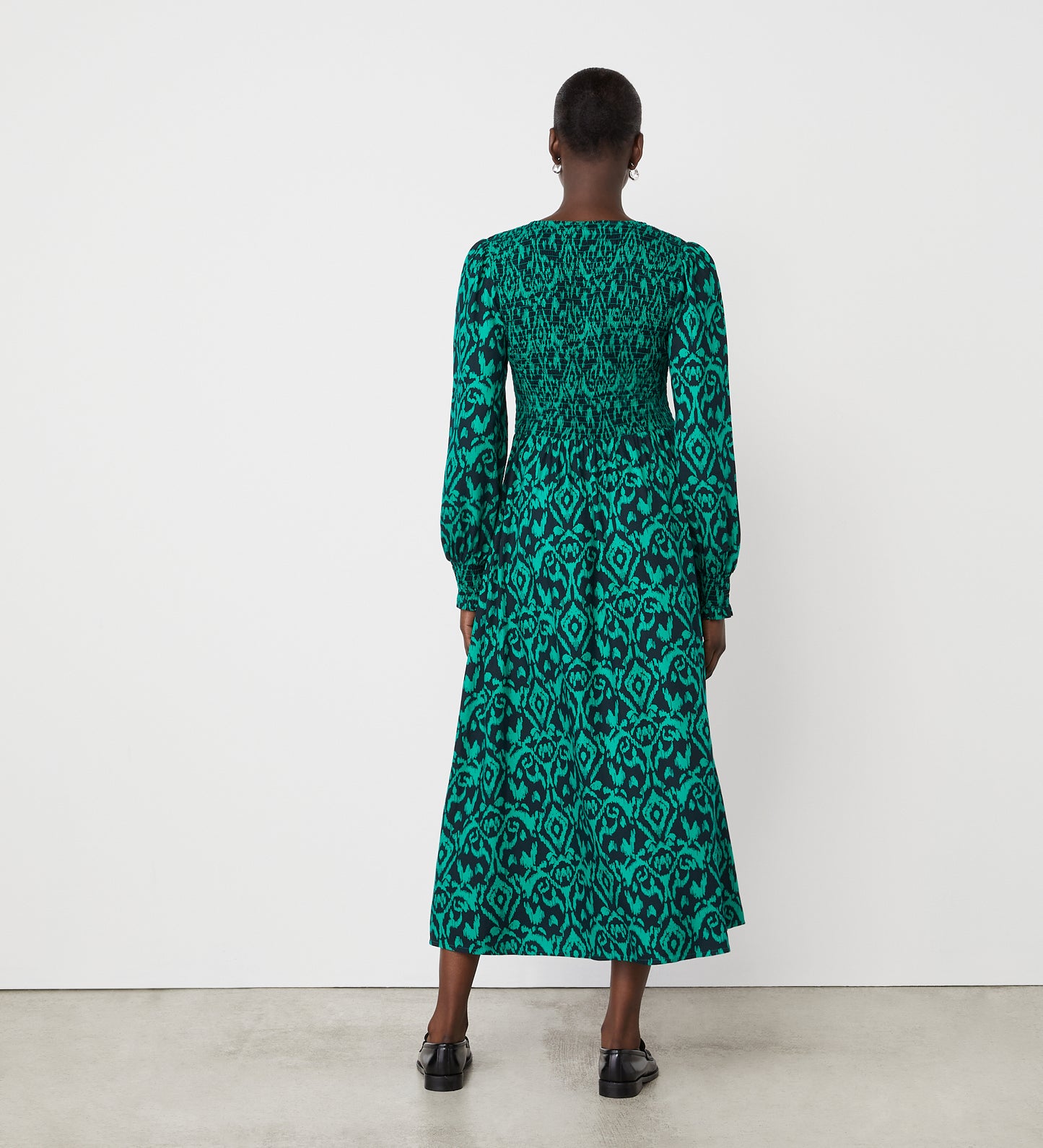 A person stands indoors on a concrete floor, facing a white wall and wearing the Tasmin Green Ikat Midi Dress—an ankle-length, long-sleeved dress with a shirred bodice and black abstract print—paired with black shoes.